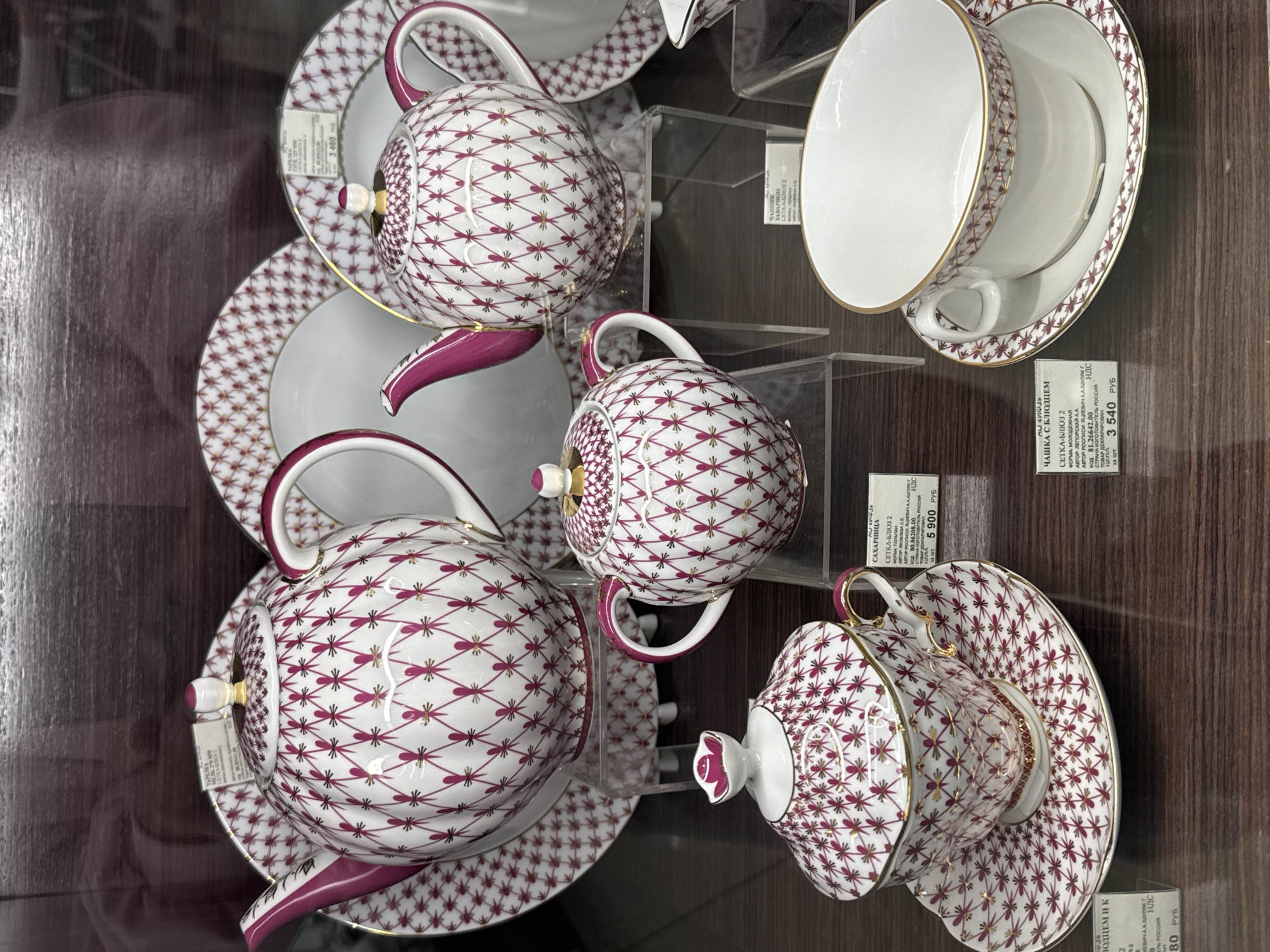The image shows a display of porcelain tea sets with a distinct pattern inside a store. The tea sets are white with a maroon and gold net-like pattern. There are teapots, cups, and saucers. Each item has a label with Russian text, including prices ranging from 3,540 to 5,900 rubles. The labels indicate the items are produced in Russia, and one item is described as being from the "Setka-Blues 2" collection, with a "Tulpan" shape. Information on the labels attributes the designs to artists named Yakovleva and Yatskevich. The photo appears to be taken at an angle on a brown shelving unit. The setting is likely a retail environment selling high-end porcelain. Given the information on the labels, these are likely tea sets from the Imperial Porcelain Factory in Saint Petersburg, Russia.