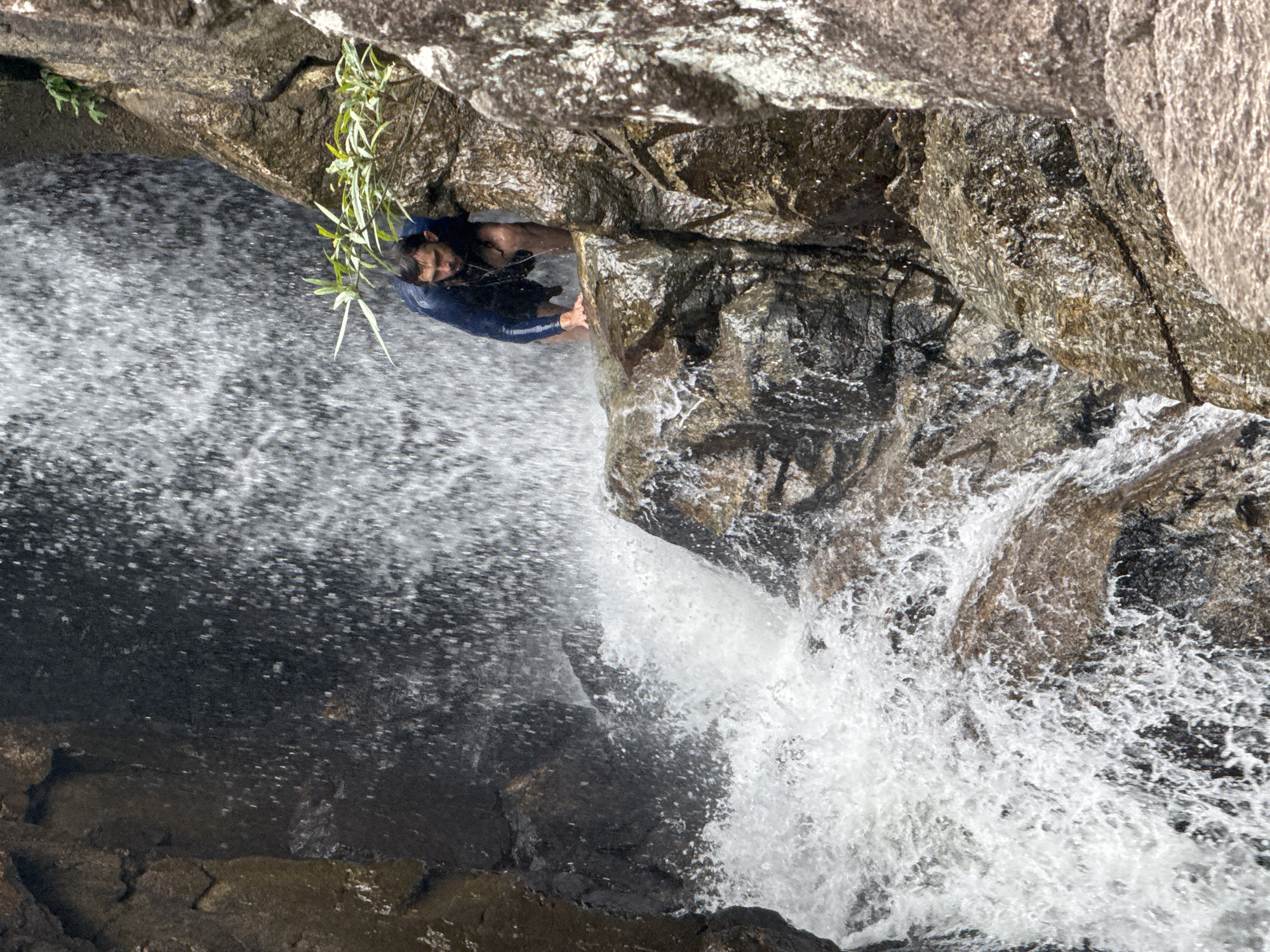 An eye-level, rotated, outdoor shot shows a man with dark hair in a dark wetsuit clinging to rocks beneath a waterfall. The man's upper torso is visible as he faces the camera. The waterfall cascades over dark rocks, creating frothy white water. Green foliage grows from the rocks near the man. The location is likely in or near the city of Đà Nẵng, Vietnam.
