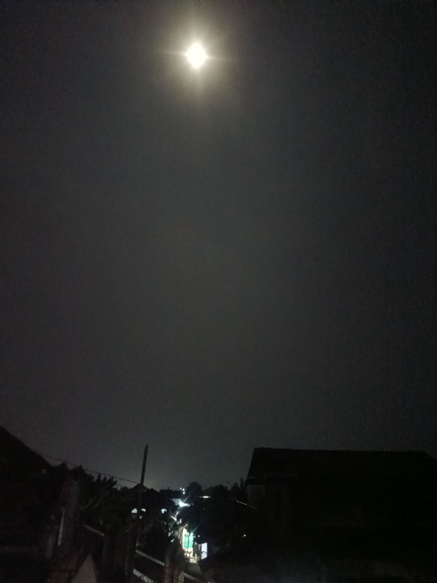 The image depicts a nighttime scene, possibly in the city of Jobokuto, Indonesia, dominated by a bright full moon in the upper center of the frame. The sky is dark and overcast with clouds partially obscuring the moon, creating a hazy glow. Below the sky, the landscape consists of silhouetted buildings and vegetation. In the foreground, a dark fence runs diagonally from the lower left to the mid-left. A portion of a dark roof is visible on the right side of the frame. Scattered lights from what appear to be buildings or streetlights are visible in the middle ground, suggesting a residential or urban area. The overall atmosphere is one of a quiet night.