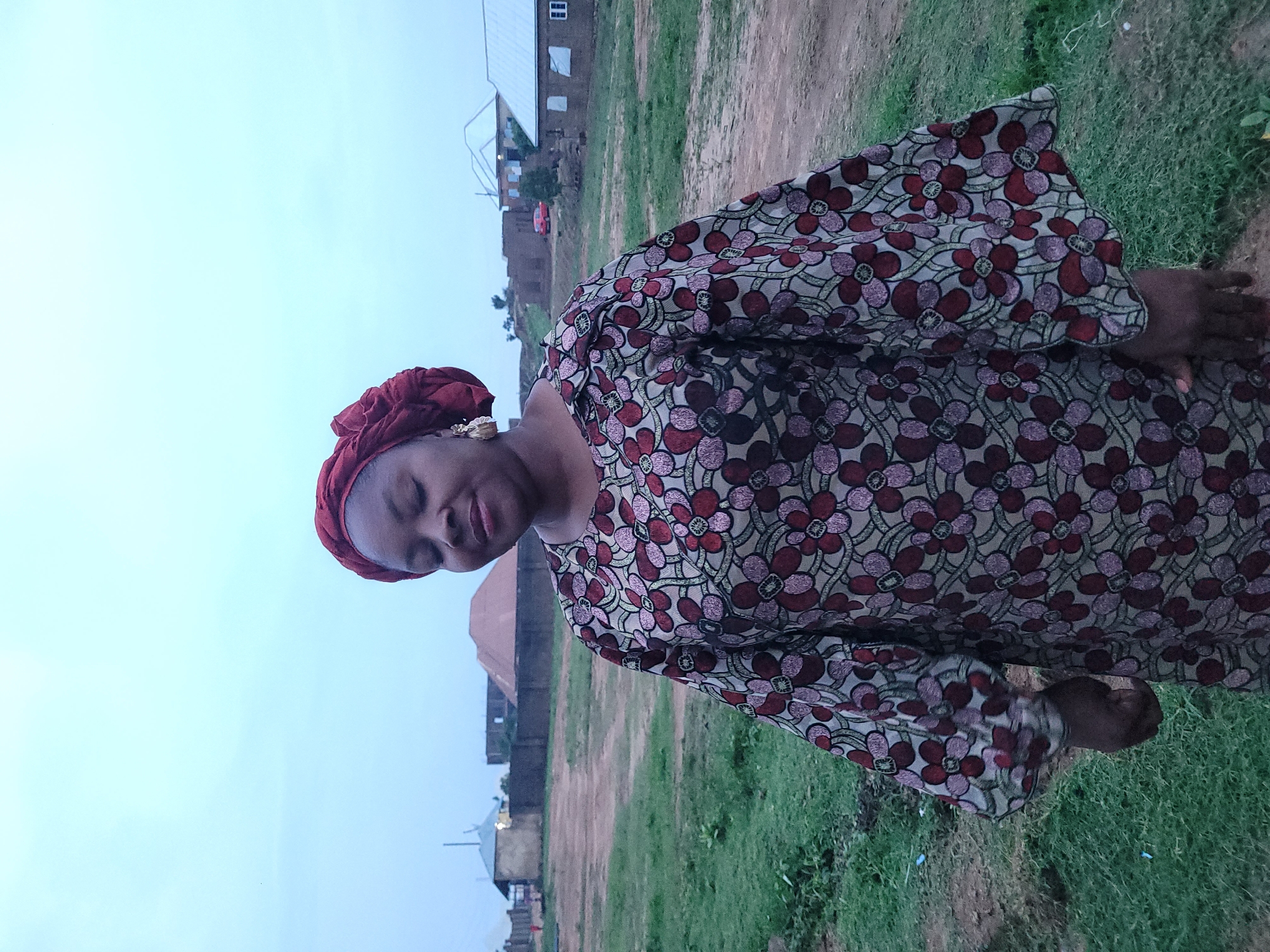 A full-shot, eye-level photograph captures a woman standing in an outdoor, semi-rural setting. The woman, positioned in the center of the frame and facing forward, wears a patterned dress with a red and floral design. She has closed eyes and her head is tilted slightly downward, suggesting a moment of repose or contemplation. Her hair is covered by a red headscarf, and she is wearing earrings. The background depicts a modest village scene with several small buildings, some appearing to be under construction, and a dirt path. A red car is visible in the middle distance. The overall environment suggests a dry, somewhat arid landscape with sparse vegetation. The sky is overcast with a pale blue hue, indicating potentially early morning or late evening light. The location is identified as Barakin Akawo, Nigeria.