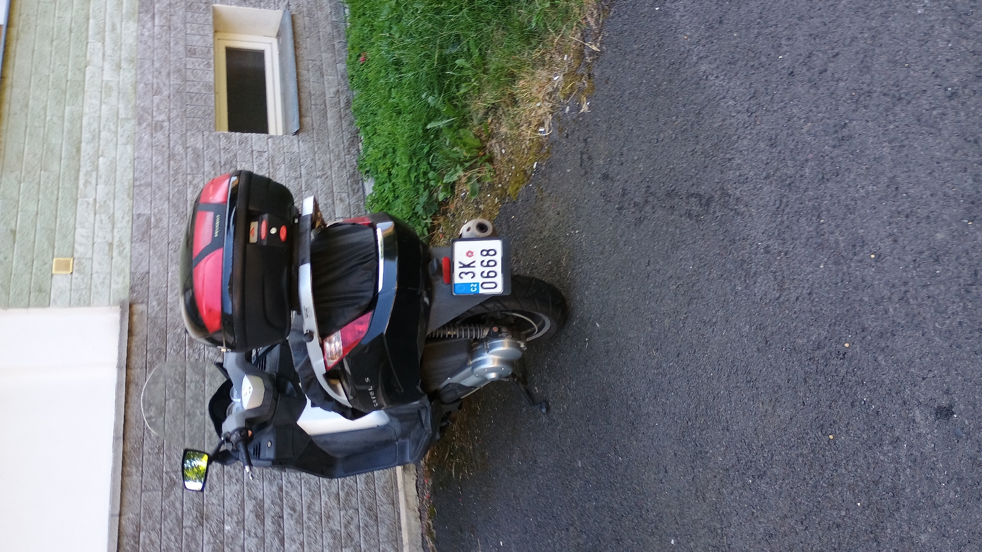 An outdoor, eye-level, vertical full shot captures a black Peugeot scooter parked on an asphalt surface. The scooter is positioned at an angle, facing towards the left of the frame. Its rear is prominently displayed, featuring a black top case with red accents, a license plate reading "3K 0668 CZ," and a silver exhaust pipe. The scooter's front is partially visible, including a reflective windshield and a black side mirror.
Behind the scooter, a gray brick wall serves as the background. The wall is partially covered by a strip of light-colored material at the top and features a small, rectangular window with a dark pane. To the right of the wall, a patch of green grass and some weeds are visible, with a brown, uneven ground surface. The overall lighting suggests a daytime scene with some shadows. No people or animals are present in the image. The setting appears to be an urban or suburban environment.