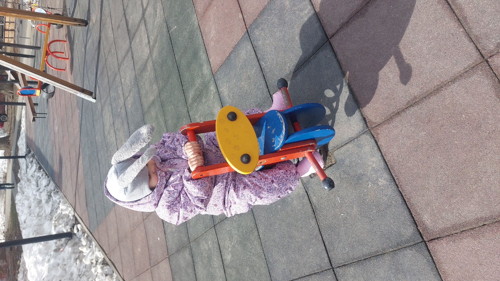 A small child wearing a hooded jacket and bunny ears is seated on a playground spring rider shaped like an airplane. The rider is constructed of red, yellow, and blue painted wood with black hand grips and foot rests. The ground is covered in interlocking rubber tiles in shades of grey, red, and brown. Remnants of snow are visible in the background. The location is in the city of Novosibirsk, Russia. Other playground equipment is partially visible in the background.
