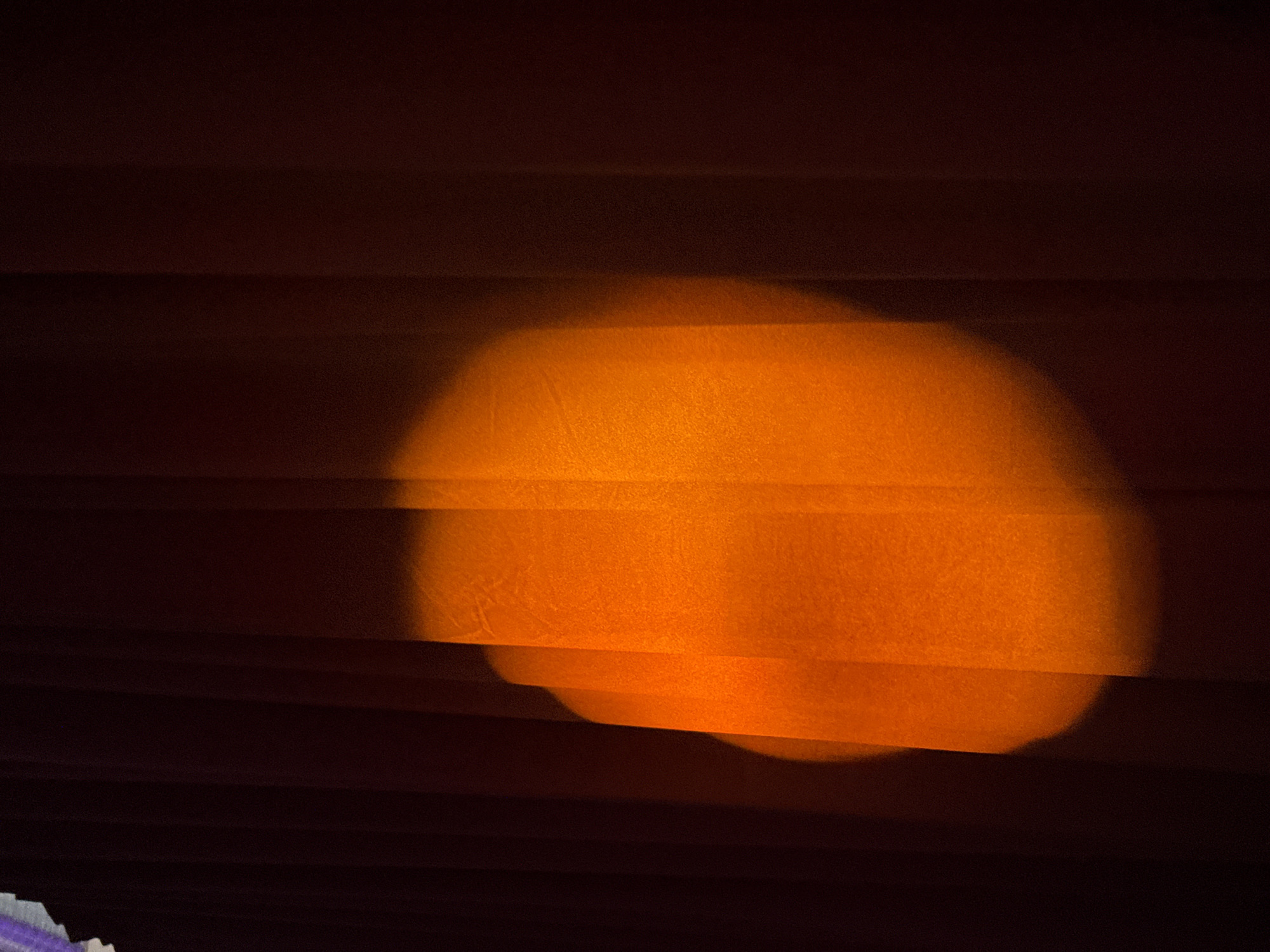 The image shows a close-up of horizontal blinds, predominantly in shades of dark brown and orange. A blurred, orange light source is visible through the blinds, creating a soft, diffused glow. There are no people or identifiable subjects present. The environment is indoors, suggested by the presence of the blinds. The location, Perm, Russia, is not evident from the visual information provided in the image.