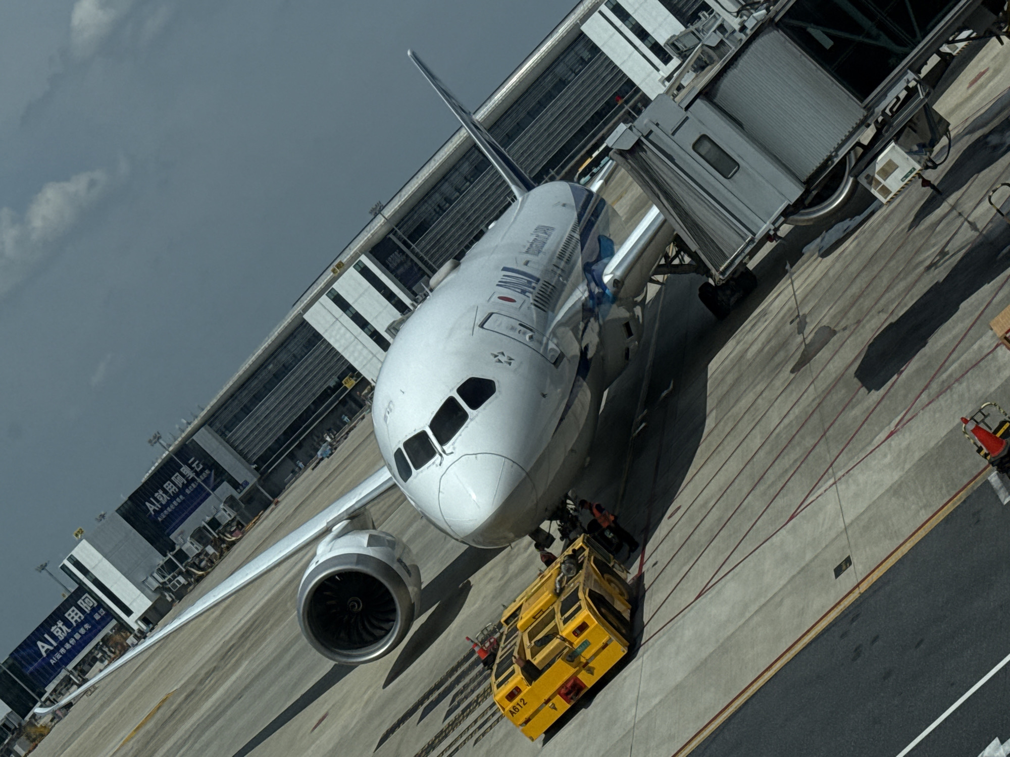An aerial, tilted view captures an airplane parked at an airport gate. The plane, a white Boeing 787 with ANA branding, is positioned at a gate, with a jet bridge connected to its fuselage. In front of the plane, a yellow baggage tug is attached to the nose gear. The airport terminal building, featuring large banners with Chinese text, is visible in the background. The scene is set on an overcast day. The airport is Hangzhou Xiaoshan International Airport, located in the Xiaoshan District of Hangzhou, China.