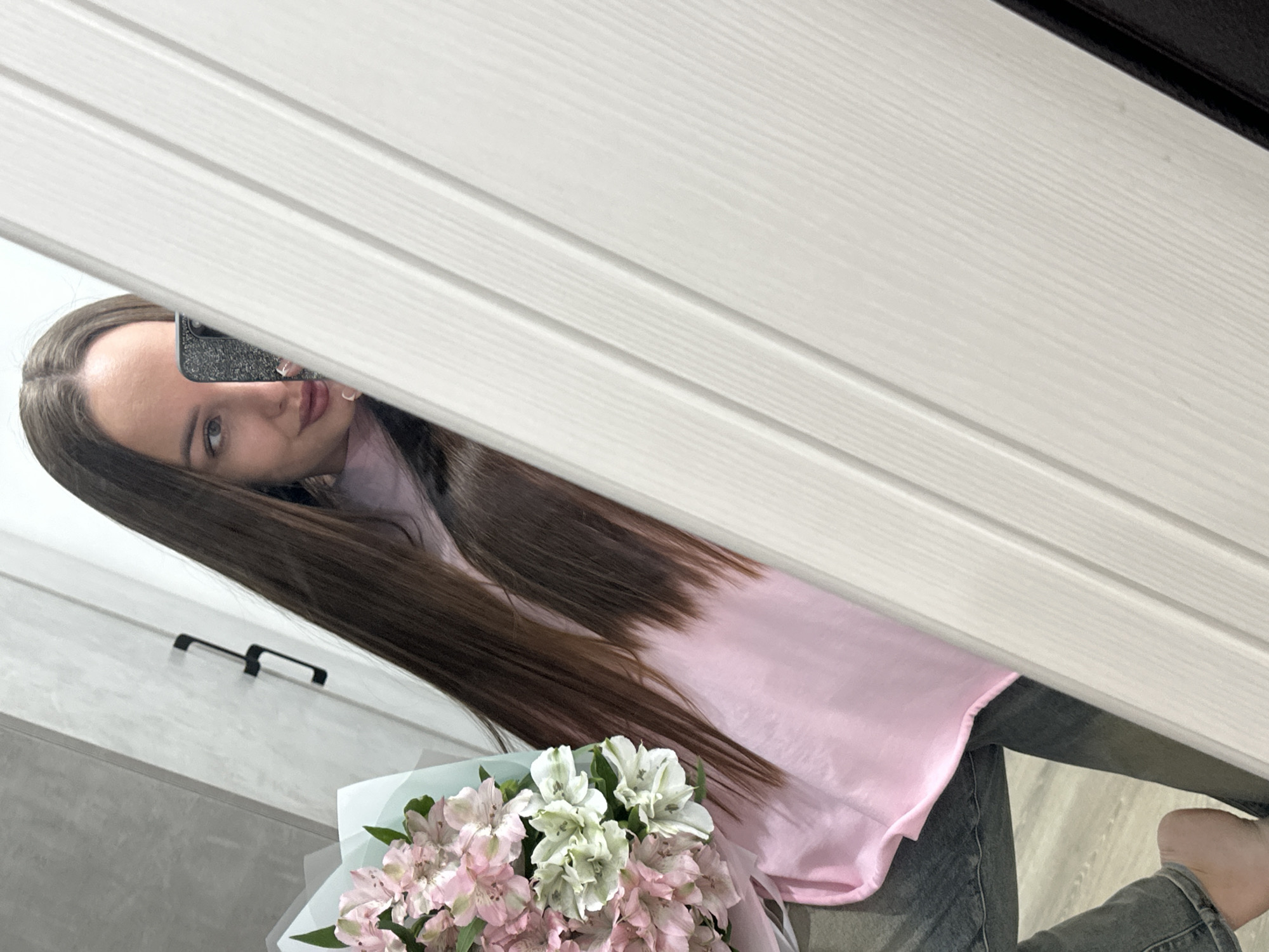 This image depicts a mirror selfie taken indoors in Chaikovsky, Russia. A female subject, with long, straight brown hair and a light pink top, is positioned center-left. She is holding a dark-colored smartphone with a textured case in her right hand, partially obscuring the lower half of her face. Her left eye, eyebrow, and lips are visible, showing a slight smile. In her left arm, she holds a bouquet of light pink and white alstroemeria flowers, wrapped in light blue and white paper. The background reflected in the mirror shows a light grey, textured wall with two black, C-shaped handles on the left. To the right, a white or off-white panel with vertical grooves, resembling a door or cabinet, occupies the majority of the right side of the frame. The subject's bare left foot and a light-colored floor, possibly wood or laminate, are visible in the bottom left corner.