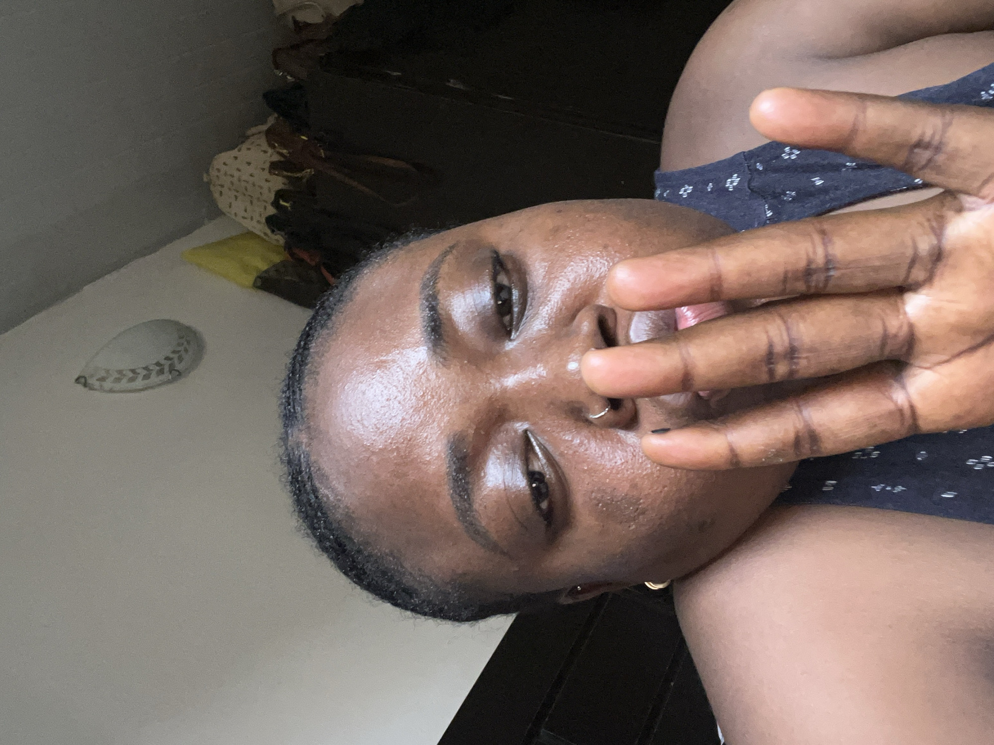 A medium-close up angled selfie depicts a woman with dark brown skin. She has short, dark hair, and her face is slightly oily or shiny. She is wearing a dark blue garment with small white floral patterns. Her left hand is raised in front of her face, partially obscuring her mouth and nose. She has a ring piercing in her left nostril, and a gold hoop earring in her left ear. A white baseball-shaped object can be seen on a white surface in the background, along with a dark wooden dresser and some purses and bags. The location is presumed to be indoors, but the specific location in Abuja, Nigeria cannot be determined from the image.