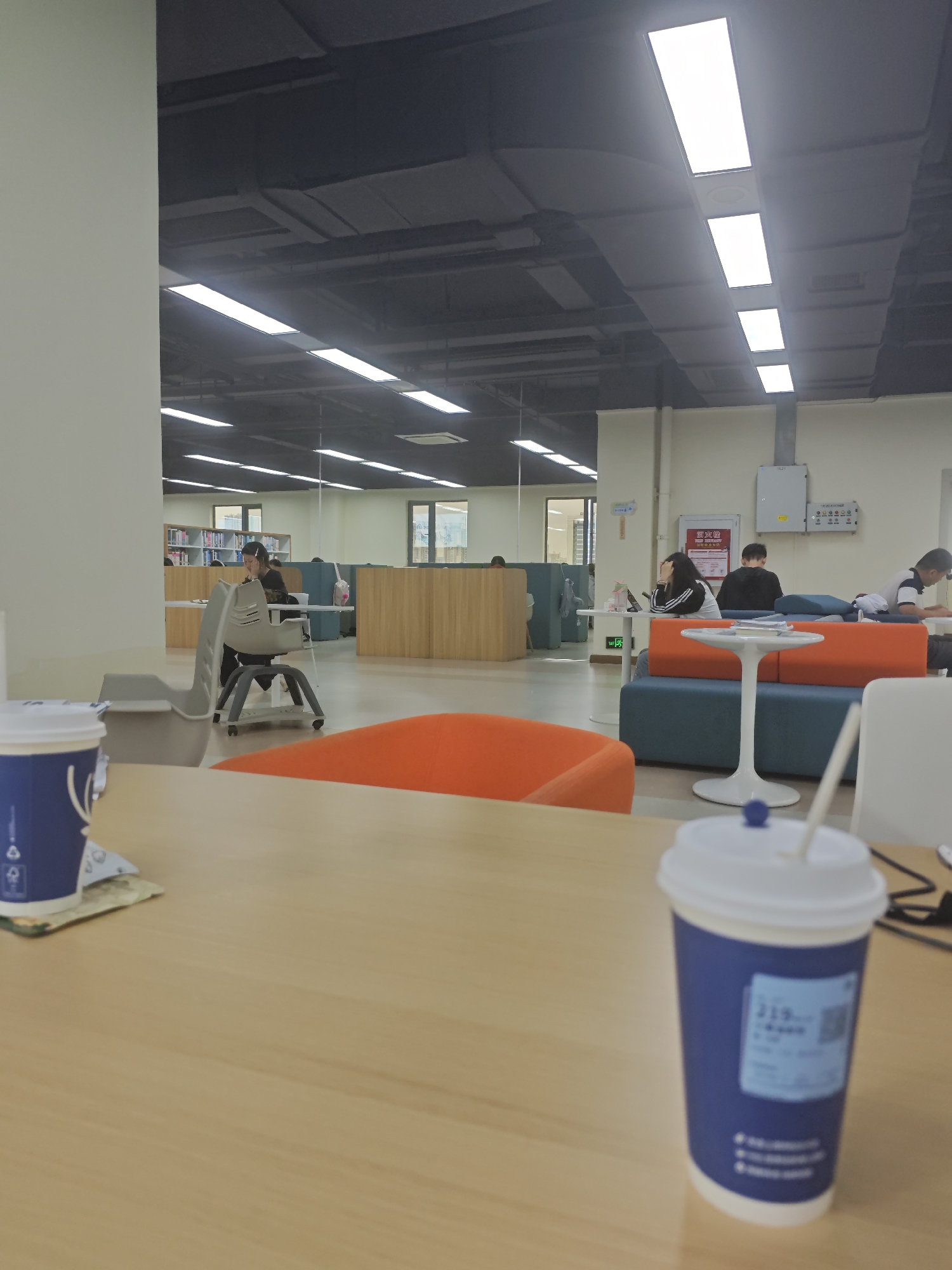 The image depicts an indoor scene, likely a library or study area, with several people engaged in individual activities. In the foreground, a wooden table is visible with two blue and white paper cups. The cup on the right is in focus, showing a white lid with a blue stopper and a straw. The cup on the left is slightly out of focus.
In the mid-ground, several seating areas are arranged. To the left, a grey chair on wheels is positioned near a table where a woman is seated, looking down at her work. Further back, a row of bookshelves is visible, with more study tables and chairs.
In the background, there are various seating arrangements with orange and teal cushions. A white pedestal table is centrally located in this area. Several individuals are seated in this section, some appearing to be studying or working on laptops. A woman with dark hair is sitting at a table with her hand on her forehead, seemingly frustrated. Next to her, a person in a black sweatshirt is looking down. To the right, a person wearing a black and white shirt is also seated.
The environment is well-lit with artificial lighting from long rectangular fixtures in the dark ceiling. The walls are a light color, and wooden partitions are used to divide the space. A white electrical panel is mounted on the wall in the background. The overall atmosphere suggests a quiet, academic setting.