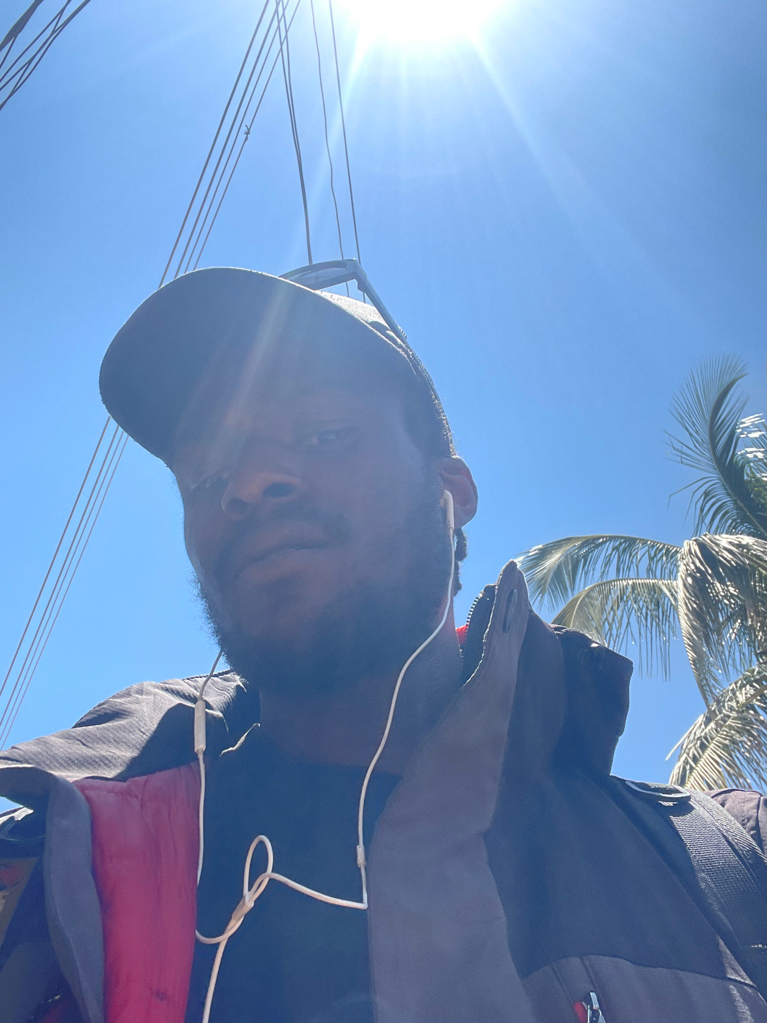 A low-angle, close-up, outdoor selfie shows a Black man wearing a baseball cap and headphones. He is looking directly at the camera. The man is wearing a black jacket over a dark shirt, with a section of bright red lining visible on the left side. The background is a bright blue sky with a strong sun glare in the upper left corner, casting rays across the image. Several thin, dark lines, possibly rigging or wires, are visible against the sky. On the right side of the frame, palm fronds from a palm tree are partially visible, suggesting a tropical or subtropical environment. The image appears to be taken on a sunny day. There is no clear indication of the city of Jos, Nigeria.