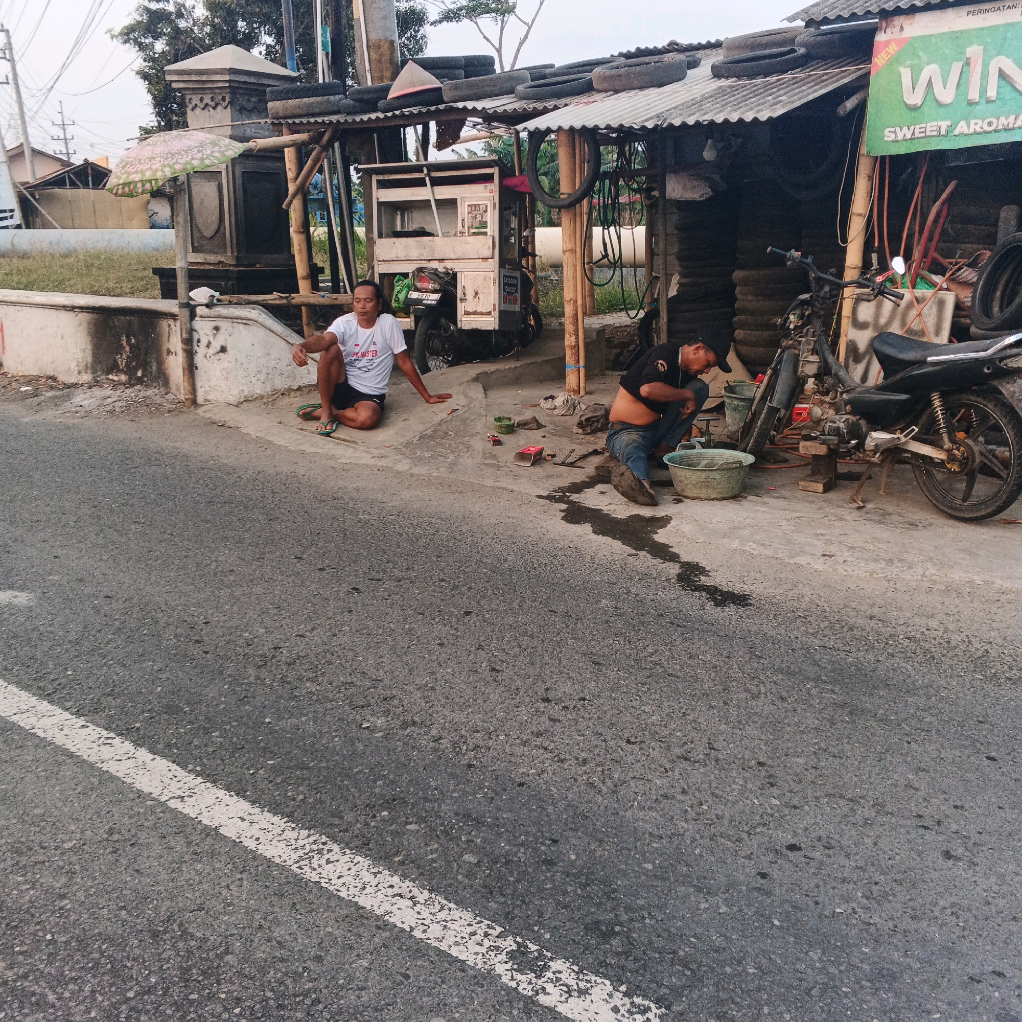 In Desa Karangkemiri, Indonesia, two men are present at what appears to be a roadside motorcycle repair or tire shop. The man on the left, wearing a white t-shirt and dark shorts, is sitting on the pavement. The man on the right, in a dark t-shirt and blue jeans, is crouched down, appearing to work on something near a motorcycle. A motorcycle is parked to the right, and a large bucket is near the crouching man. Various tires are stacked and visible, particularly on the roof of the makeshift structure behind the men. The structure itself is made of corrugated metal and wood. A sign with green and white lettering is hanging on the structure. The scene is situated beside a paved road with a white line marking.