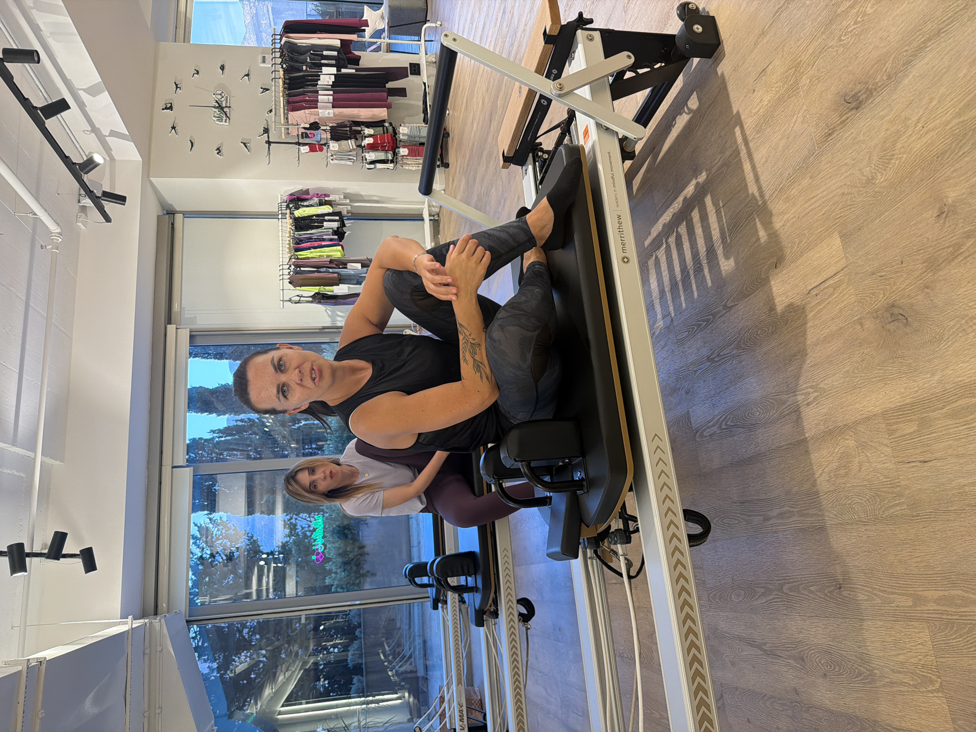 The image shows two women in a Pilates studio in Boreti, Montenegro. The woman in the foreground is sitting on a Pilates reformer, wearing a black tank top and patterned leggings. She has dark hair and tattoos on her arm. The woman in the background is sitting on another Pilates reformer, wearing a light purple t-shirt and dark leggings. The studio has large windows that look out onto trees and mountains. There are racks of athletic clothing visible in the background. The floor is made of light wood.