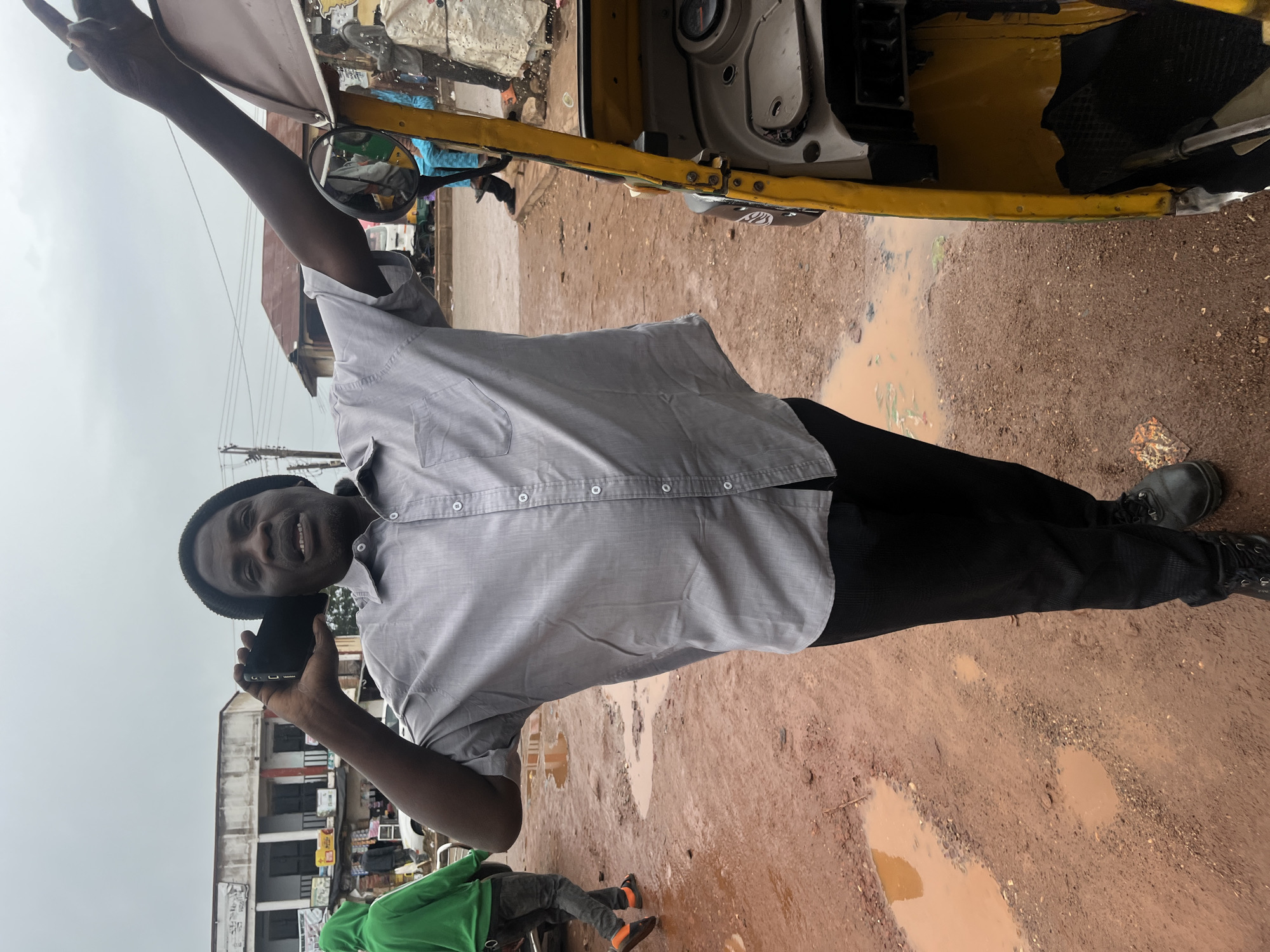 A man in a light gray button-down shirt and black pants is standing next to a yellow auto-rickshaw, holding a black smartphone to his ear and smiling. He wears a dark beanie. His left arm is raised, and he appears to be gesturing with his right hand. The scene is outdoors, with a muddy, wet ground, suggesting it may have recently rained. Several puddles are visible. In the background, there are buildings that appear to be shops or residences, with some people and vehicles present. The sky is overcast. The overall impression is a casual, everyday moment captured on a cloudy day in what appears to be a busy, informal marketplace or street setting in Bukuru, Nigeria. There is no visible text in the image.