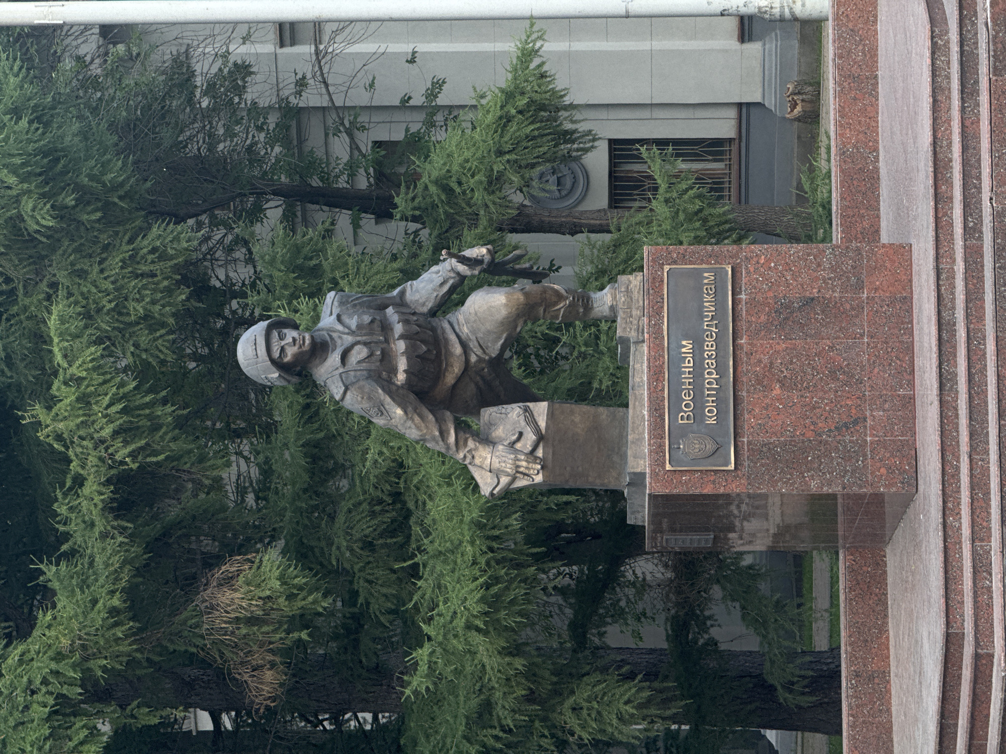 A bronze statue of a male soldier in a helmet and tactical gear stands on a granite pedestal. The soldier is depicted in a forward-leaning stance, with his left hand resting on a book or document atop a smaller block, and his right hand holding a rifle. The pedestal has a plaque with Russian text that reads "Военным контрразведчикам" (To Military Counterintelligence Officers). The statue is located in front of a building with a light-colored facade and is surrounded by lush green trees. The ground in front of the pedestal is paved with stone. This monument is located in Yekaterinburg, Russia, on the territory of the FSB Directorate for Sverdlovsk Oblast.