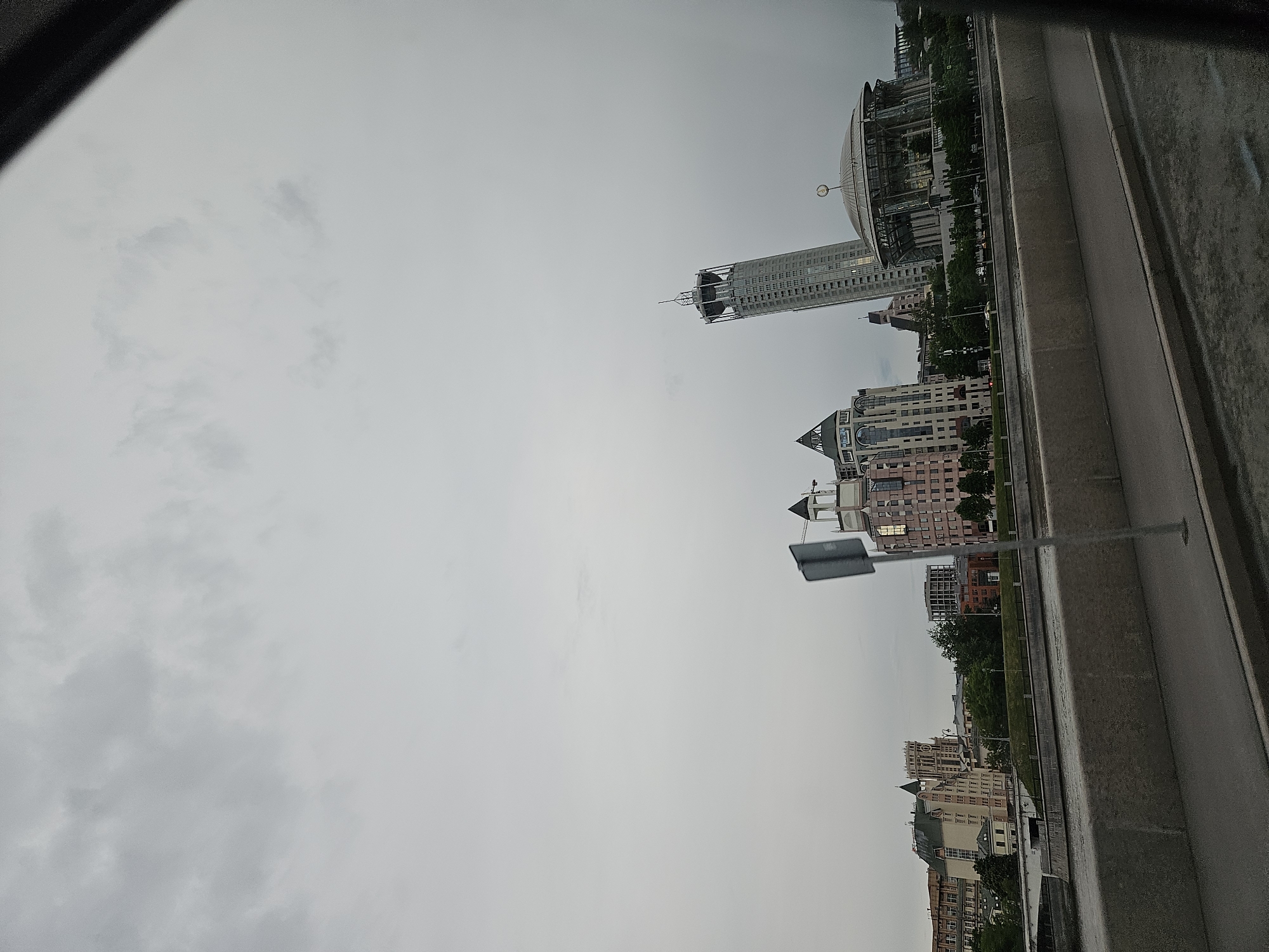 The image shows a view of the city of Moscow, Russia from an elevated position, possibly a bridge or higher roadway. Several buildings are visible, including a tall skyscraper with a distinctive dome-shaped structure atop, and several other buildings of varied architectural styles. A gray, overcast sky dominates the scene. A portion of the bridge's or roadway's barrier is visible, along with a sign. There are no people visible in the image.