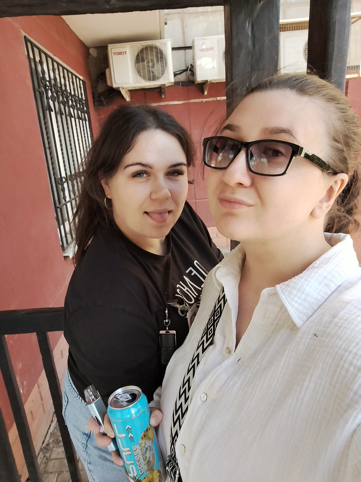 The image shows two young women standing outdoors. The woman on the left has dark brown hair and is wearing a black t-shirt and blue jeans. She is holding a vape pen in her left hand and a can of what appears to be a beverage in her right. She is sticking her tongue out and looking at the camera. The woman on the right has her hair pulled back and is wearing a white button-down shirt. She has on large, black-rimmed glasses and is also looking at the camera. The background features a red building, a barred window, and two air conditioning units mounted on the wall. The image is a selfie, with the camera held by one of the women. The scene is likely a casual outdoor setting. There is no indication of the location being in the city of Chaikovsky, Russia.