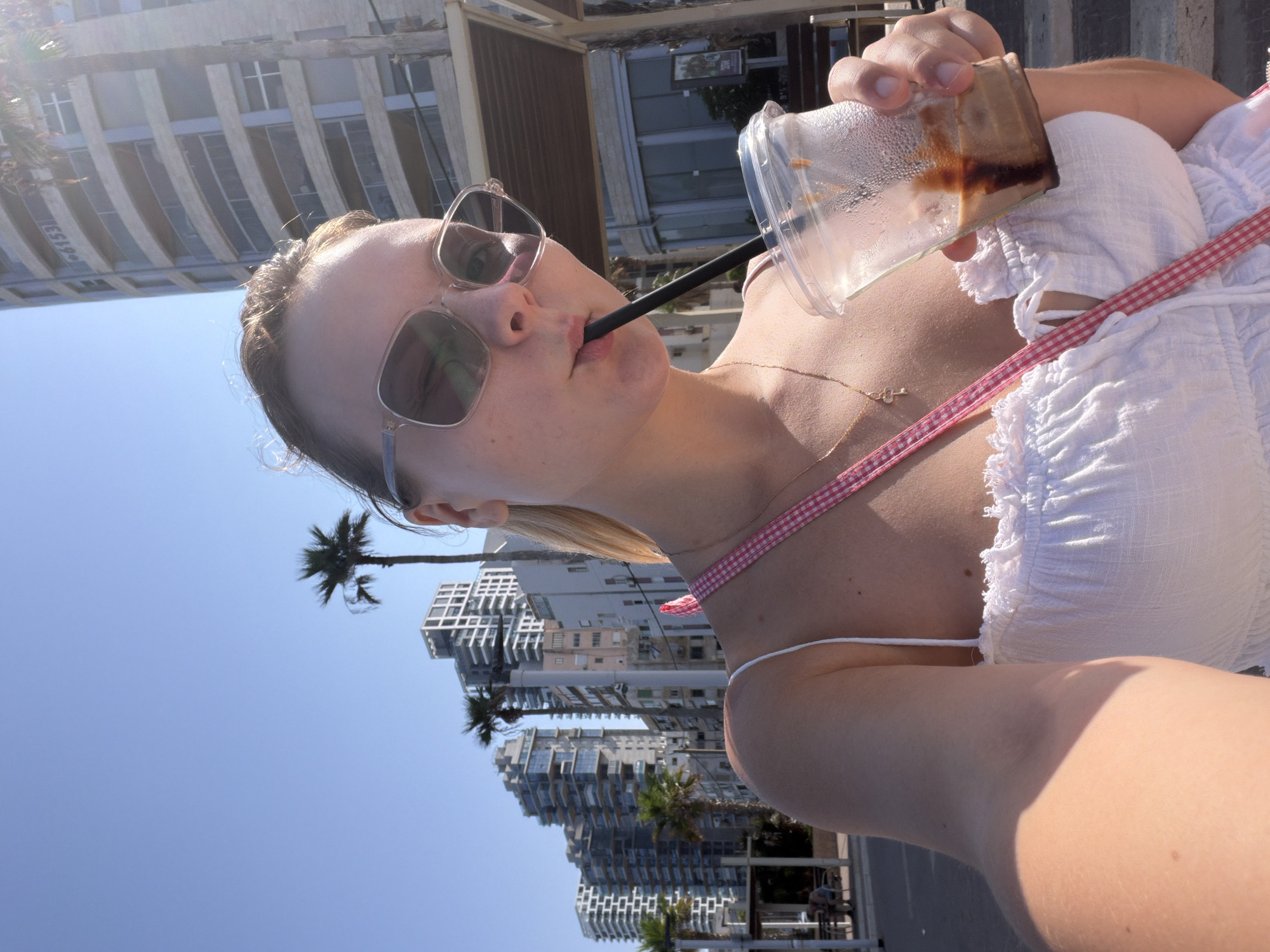 A woman with blonde hair and sunglasses drinks from a to-go cup with a straw. She is wearing a white top with a pink and white checkered strap across her chest, and a gold necklace with a key pendant. The background shows a sunny street scene in Bat Yam, Israel, with palm trees and modern buildings. The architecture suggests a coastal city, possibly near a promenade or beach access. The lighting indicates it is daytime, with the sun casting a bright glare in the upper right portion of the image.