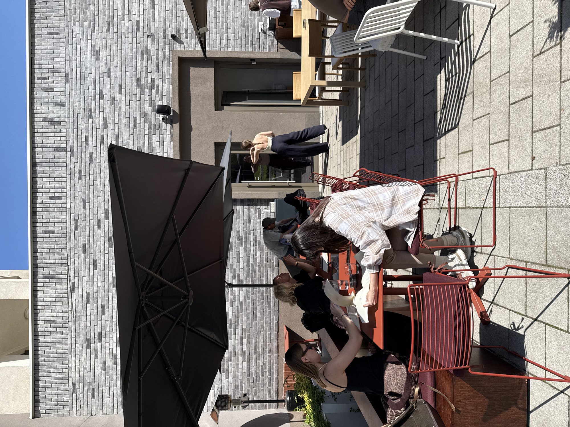 This media file captures a lively outdoor patio scene, likely at a cafe or restaurant, in Belgrade, Serbia. The image depicts a bright, sunny day under a clear blue sky, casting strong shadows from furniture onto the paved ground.
A group of people, primarily young adults, are gathered on the patio. Several individuals are seated at tables in the foreground, characterized by red metal chairs and tables. One woman in a plaid shirt and another in a black top are seated, seemingly engaged in conversation or looking at menus, though no text is visible. A large black patio umbrella on the left provides shade. In the mid-ground, a person wearing a light top and dark pants stands near a window, while other patrons are visible seated at wooden tables further back.
The setting includes a modern building with a distinctive grey and white brick facade, complemented by plain concrete sections, windows, and doors. The ground is paved with light grey rectangular tiles. The overall ambiance suggests a relaxed and casual social gathering enjoying the pleasant daytime weather.