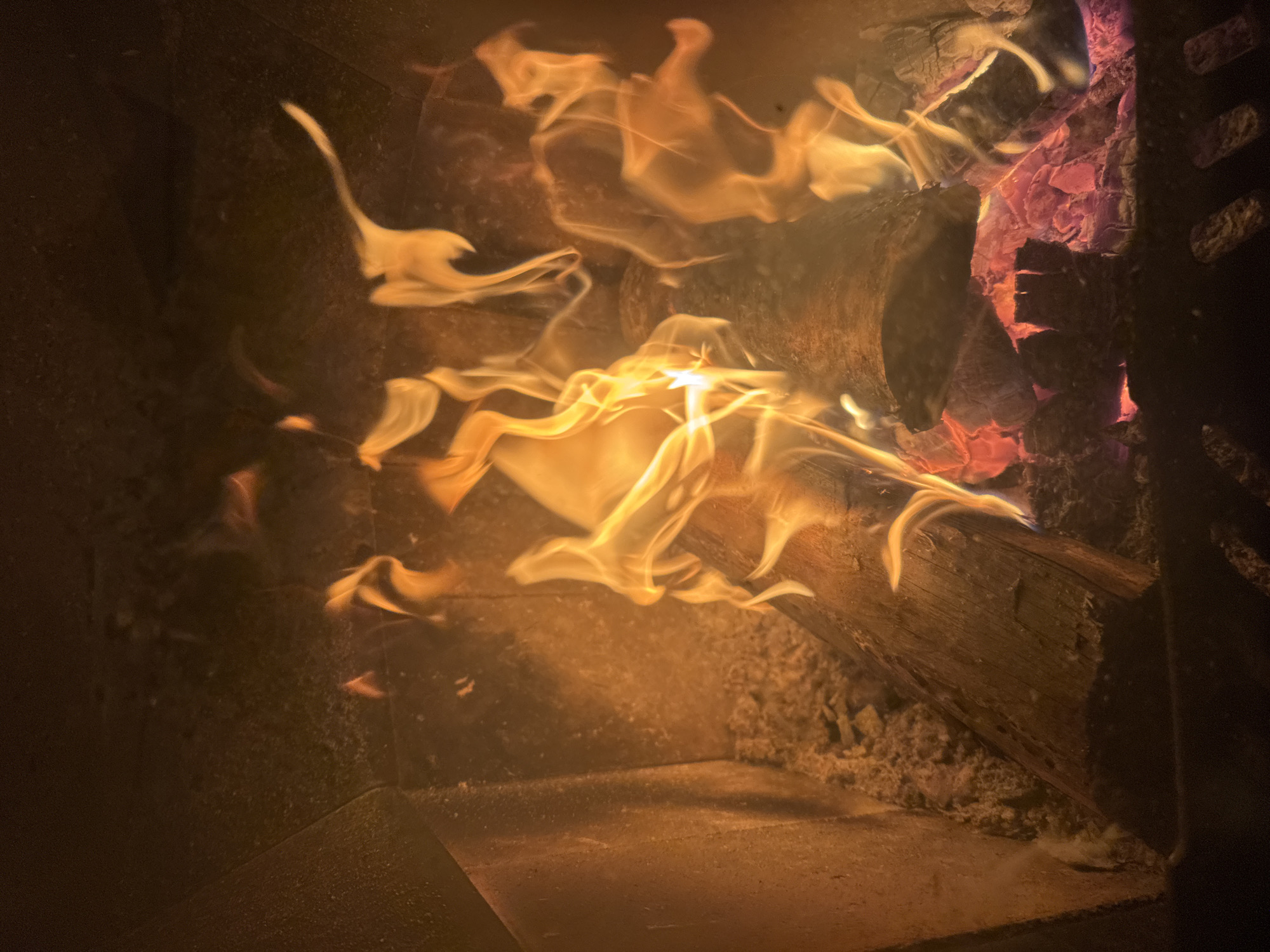 The image depicts a fire burning inside a fireplace or wood-burning stove. The flames are prominent and appear to be consuming pieces of wood. Red embers are visible at the right side of the frame. The interior of the fireplace/stove shows signs of wear and soot accumulation. The location in Mayak, Russia is irrelevant, as the scene is generic and not indicative of any specific information.