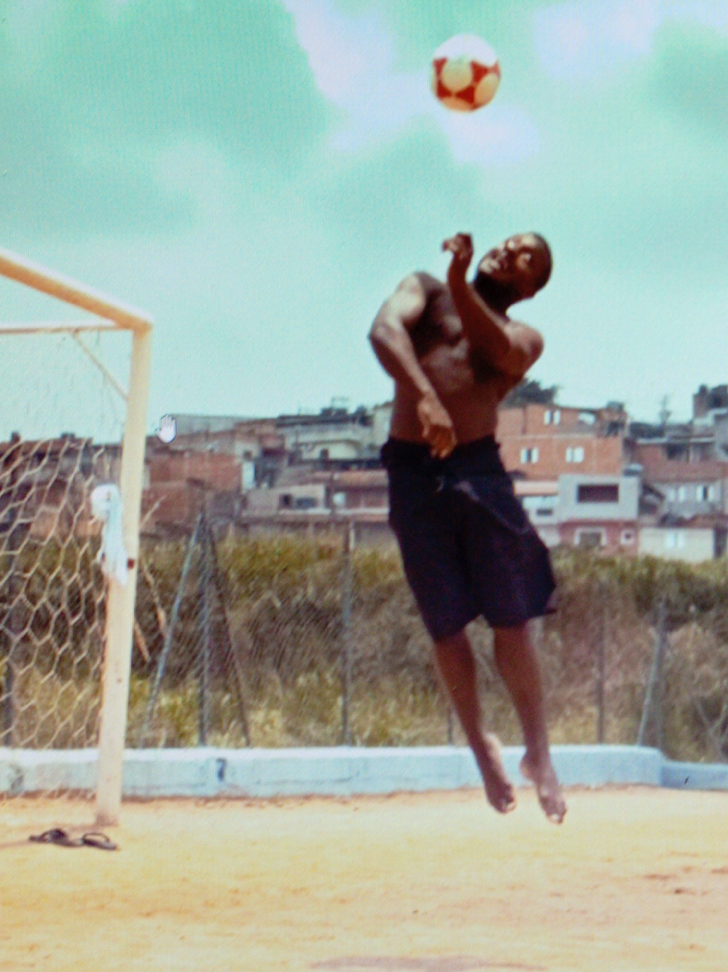 A shirtless Black man is captured mid-air, leaping to head a soccer ball. He is wearing dark shorts. The soccer ball is red and white, suspended in the bright, hazy sky. He is positioned in front of a soccer goal with a net. In the background, there are buildings that appear to be in an urban or semi-urban setting, with some vegetation visible. The ground is sandy and appears to be a soccer field. There are a pair of flip-flops on the ground near the goal post. The sky is a light blue with some soft clouds, suggesting a daytime setting, possibly with some haze or light cloud cover. The overall mood is athletic and active, focusing on the dynamic movement of the player and the ball. No visible text is present in the image.