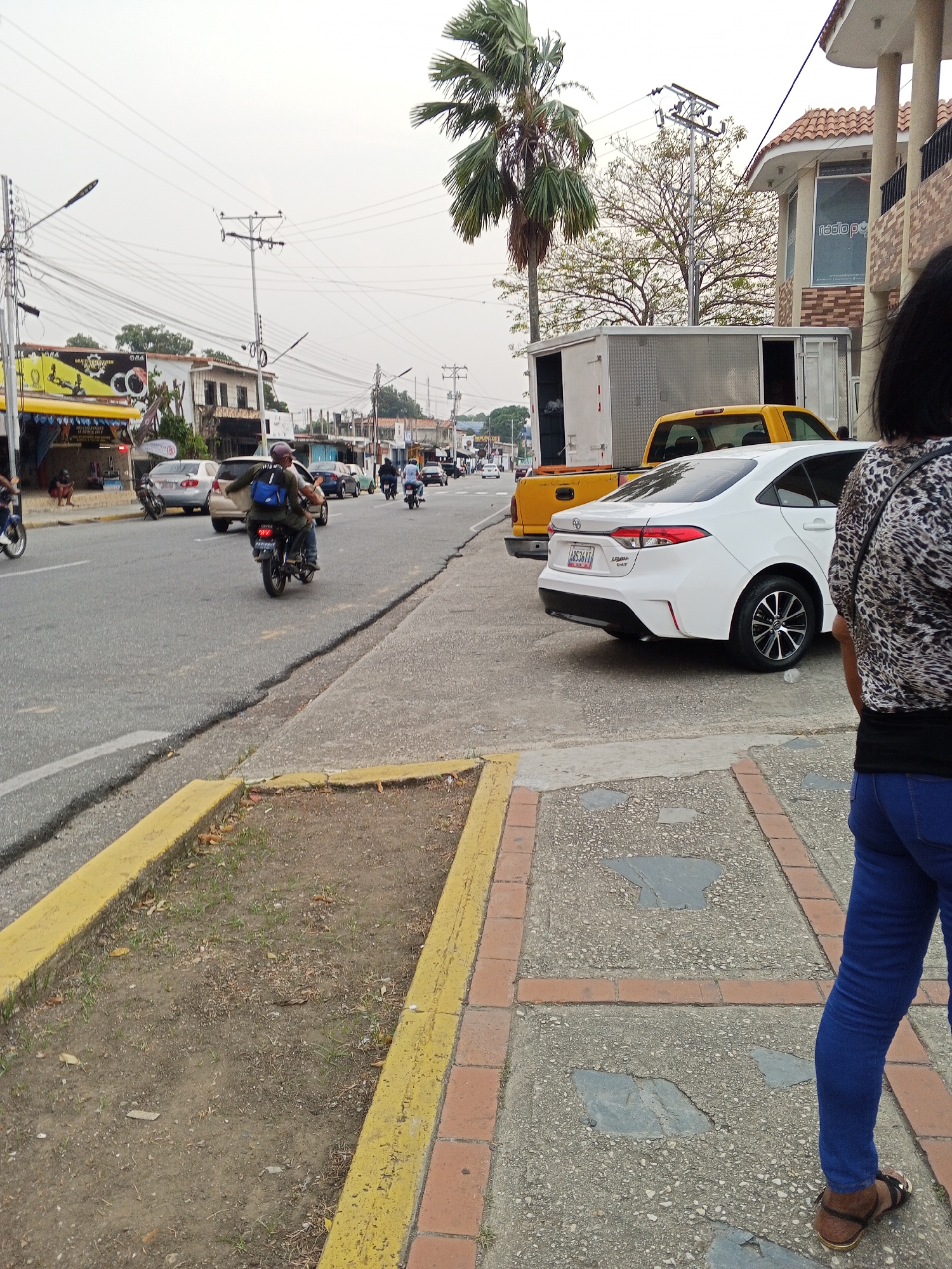 The image depicts an urban street scene in Tinaquillo, Venezuela, observed from a low angle on a sidewalk. In the immediate foreground, the back of a person wearing a leopard-print top, blue jeans, and brown sandals is partially visible on the right. A concrete sidewalk with embedded square slate-like tiles and intermittent red brick sections extends leftwards. Adjacent to the sidewalk on the left is a planter strip with yellow painted curbs, containing dry soil and sparse green vegetation.
The midground features a two-lane asphalt road exhibiting multiple cracks. Parked on the right side of the road, facing away from the viewer, is a white Toyota Corolla sedan with Venezuelan license plate A8536Y-1. Immediately in front of the sedan is a yellow pickup truck with an attached white utility box, its rear doors open. Several vehicles are in motion on the road, including multiple motorcycles. One motorcyclist, wearing a helmet and a blue backpack, is prominently visible in the left midground, traveling away from the camera. Other cars are visible further down the street in traffic.
Along both sides of the street, commercial and residential buildings line the roadway. On the left, several single and multi-story buildings house storefronts with visible signage, including one yellow awning with "ORA" text and displays featuring electronic equipment like ring lights. A person is observed sitting on a low wall near this storefront. On the right, a multi-story building with a red tiled roof and columns features a large window with "radopop" text. Overhanging power lines and utility poles are abundant throughout the scene. A tall palm tree stands on the right, and a deciduous tree with sparse foliage is visible behind the parked vehicles. The sky is overcast, indicating a cloudy day.