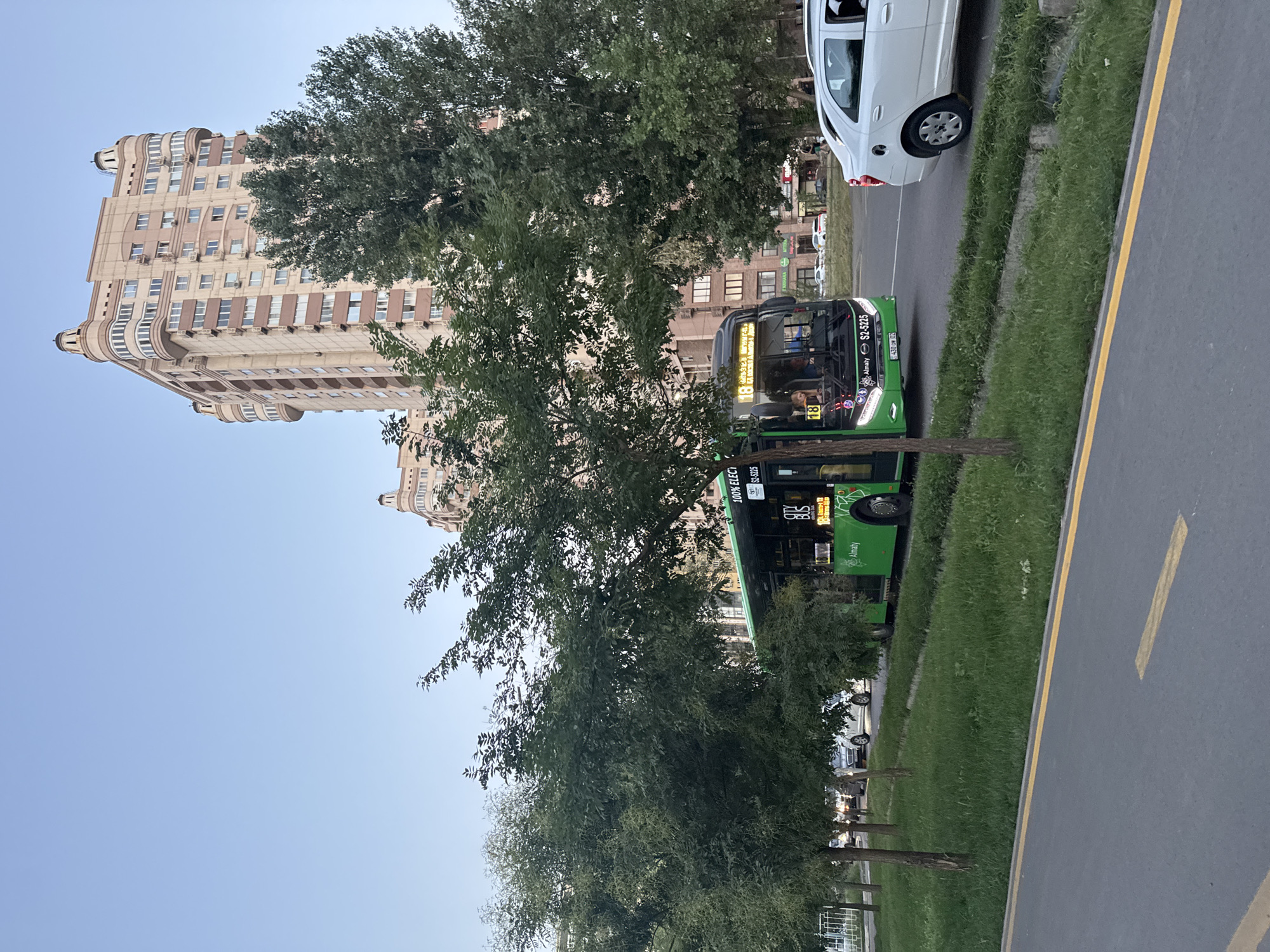 The image depicts a street scene in Almaty, Kazakhstan, under a clear, pale blue sky. A modern, green city bus, identified with the number "18" and "CITY BUS", is the central subject, traveling on the road. The bus displays "100% ELECTRIC" on its side and the Almaty logo. To the right of the bus, a white car is also in motion. In the background, a tall, light-colored residential building with distinctive architectural details, including rounded corner balconies and ornate top sections, dominates the skyline. Several trees with dense green foliage line the street, partially obscuring the view of the buildings. The foreground shows a paved road with yellow lane markings and a strip of green grass. Several other vehicles and a portion of another apartment building are visible further down the street.