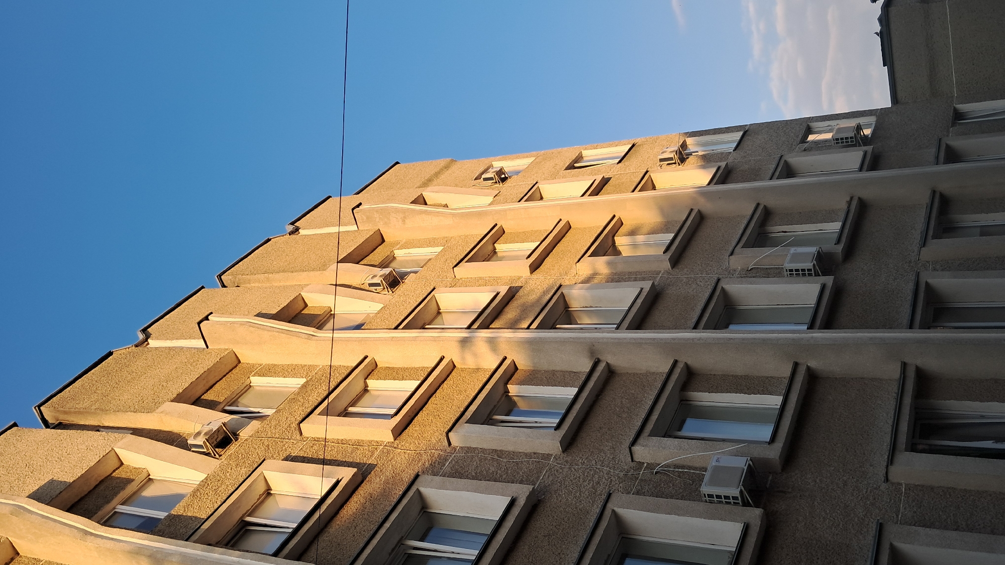 The image displays the exterior of a multi-story building, likely a residential or office complex, under a clear blue sky. The building's facade is characterized by a textured, light brown or beige material and features a repeating pattern of windows and vertical architectural elements. Several windows are visible on each level, some appearing to be open, revealing the interior. Air conditioning units are mounted below some of the windows. The building is illuminated by sunlight, casting long shadows that accentuate its geometric design. A thin wire stretches horizontally across the upper portion of the image. The lower part of the image shows a glimpse of another building with a dark roof. The scene is devoid of people.