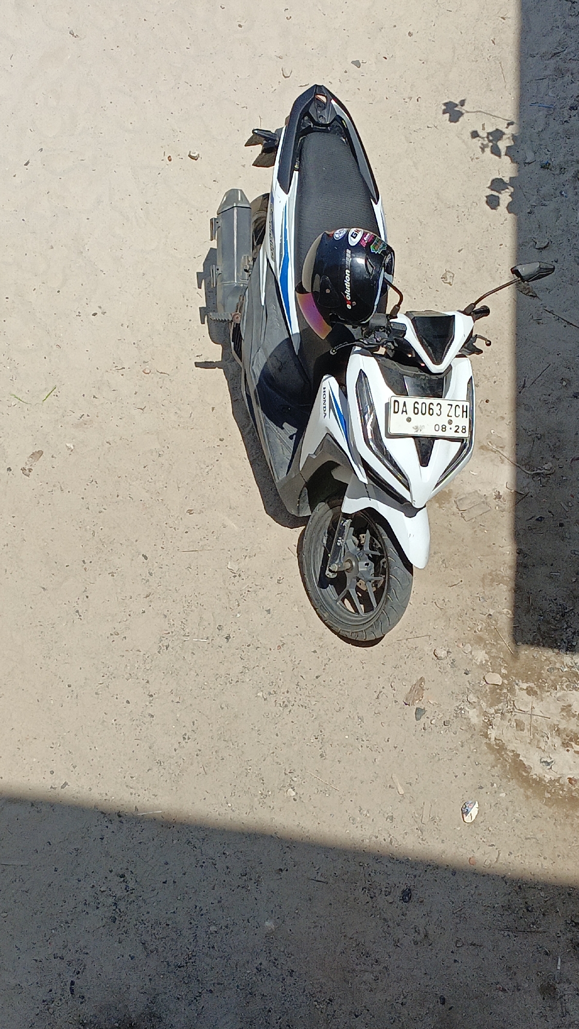 A black and white scooter is parked on a dirt ground. A black helmet with some stickers is placed on the seat of the scooter. The license plate on the front of the scooter reads "DA 6063 ZCH 08-28". The ground is uneven and speckled with small rocks and debris. A shadow from a tree falls across the ground and the scooter. The image appears to be taken from an elevated position looking down.