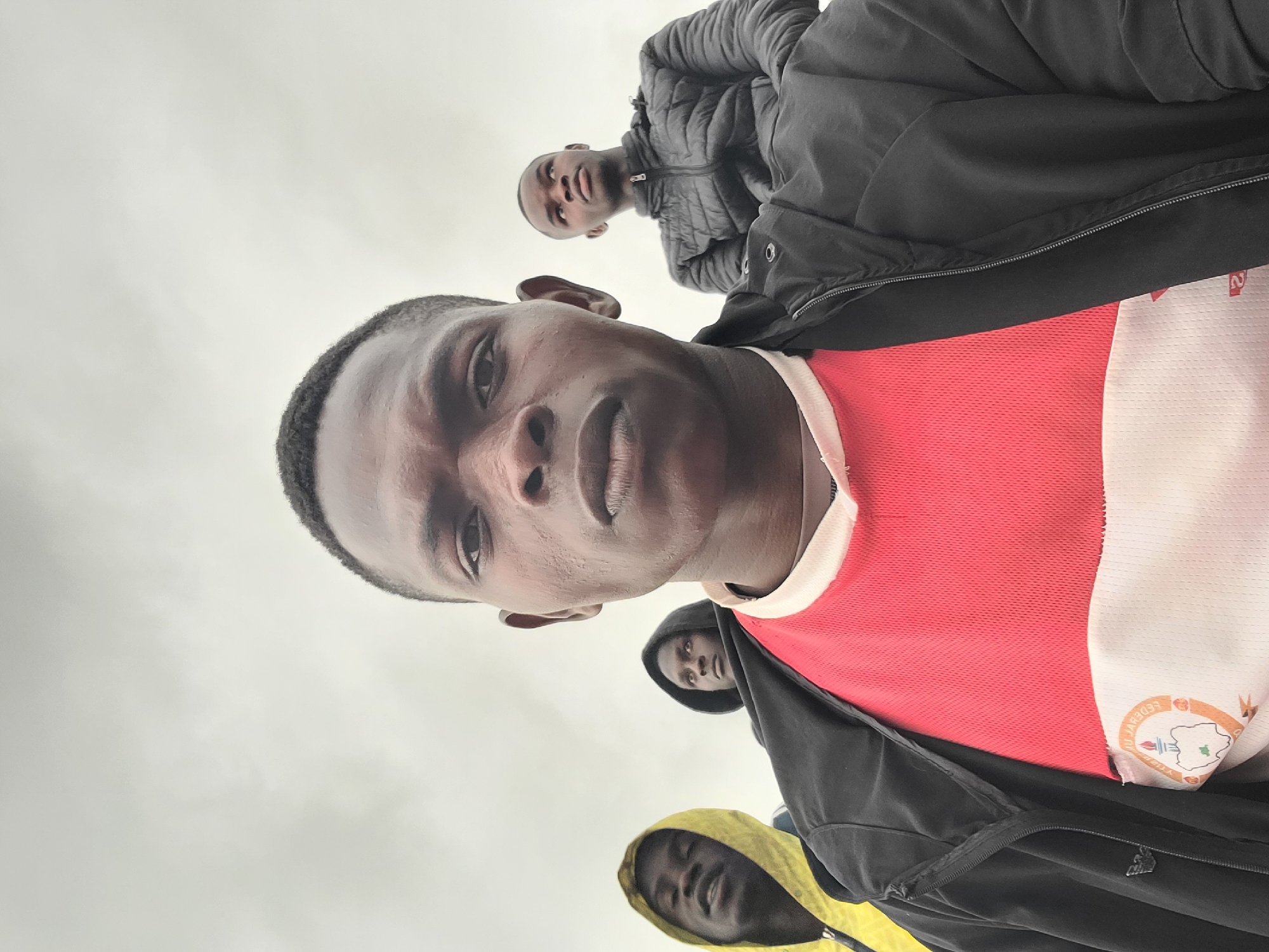 The image depicts four young Black males captured from an extreme low-angle perspective, looking up towards them against a bright, uniformly light sky, suggesting an overcast day.
In the immediate foreground, a young man with short, dark hair and a serious or neutral expression looks directly at the camera. He is wearing a red short-sleeved top with a white collar, and a black zippered jacket partially covers it. On the white section of his top, near his right shoulder, a circular badge or logo is visible. The text on the badge is partially obscured and mirrored due to its placement, but fragments like "FEDERB" and a geographical outline or emblem can be discerned.
Behind him and slightly to his left, another young man is visible from the chest up, also looking towards the camera with a neutral expression. He wears a black puffer jacket. Further back and below these two, two other young men are partially visible. One, centrally located in the background, wears a dark hooded jacket with the hood up, looking upwards. To his left and slightly lower, another young man in a bright yellow hooded jacket, also with his hood up, appears to be looking up with a relaxed or slightly smiling expression.
The plain, bright sky serves as the only background, providing no specific location cues. The diffused lighting suggests it is daytime. The image appears to be a group portrait or selfie taken from a very low camera angle, with the subjects looking down towards the lens.