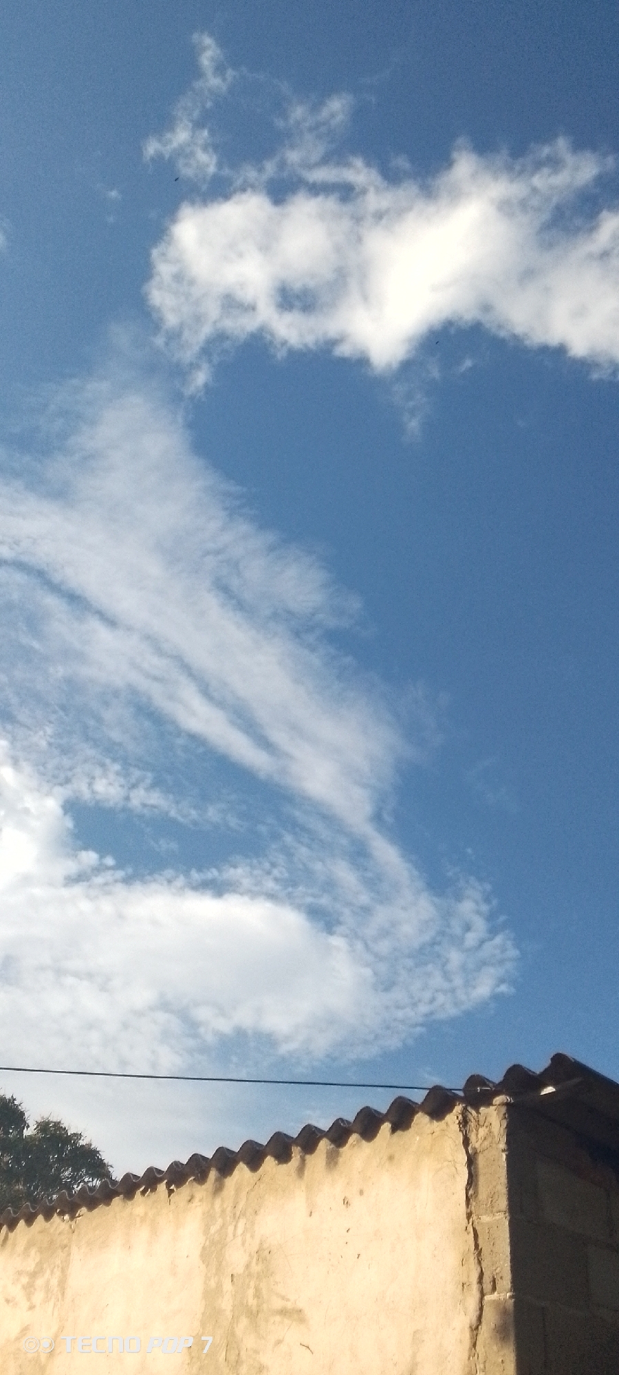The image shows a view of a blue sky with white clouds above a building in Altagracia, Venezuela. The building has a corrugated roof and a textured, light-colored wall. A few tree leaves are visible to the left of the building. A power line crosses in front of the roof. The sky contains both cumulus and cirrus clouds.