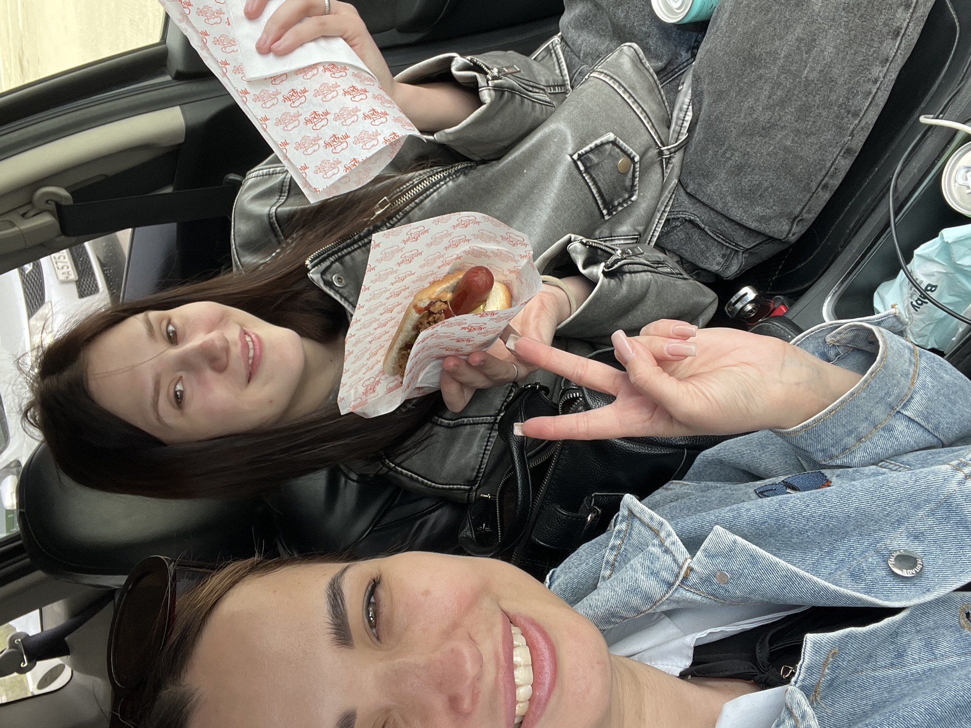 Two young women are in a car. The top woman is holding a hot dog and a paper wrapper with a red design. The woman at the bottom of the image is giving a peace sign and smiling. The women are wearing jackets. The top woman is wearing a gray leather jacket, and the bottom woman is wearing a blue denim jacket. There are cans in the background and a baby wipe container. The location is unknown.