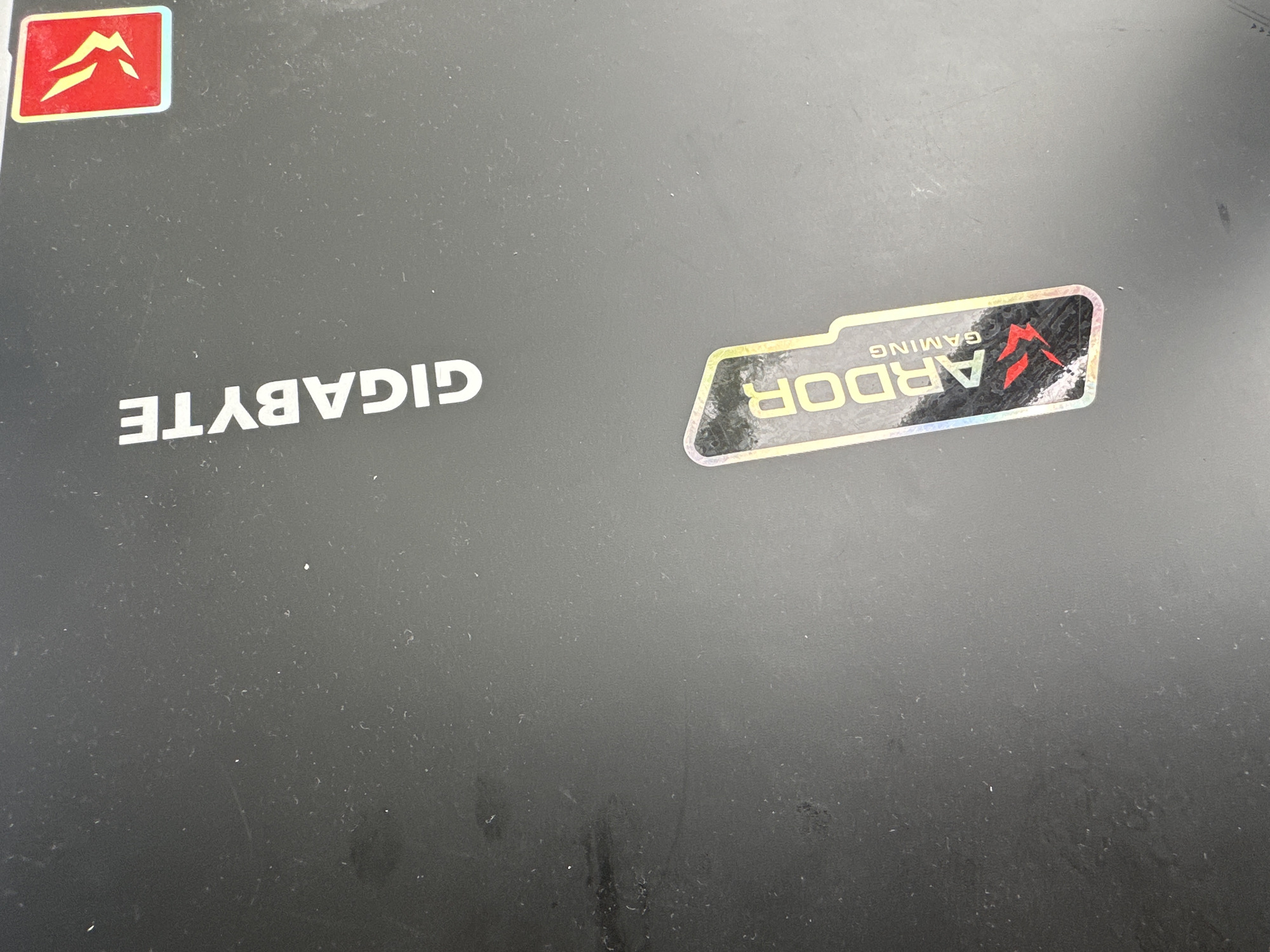 The image shows a black surface, likely a laptop lid, with two stickers. On the left, a red and yellow sticker features a mountain-like symbol. On the right, a holographic sticker displays the word "ARDOR" in large letters, with "GAMING" written above it in smaller text. The surface appears dusty and has some smudges. The location is not visible in the image and cannot be determined.