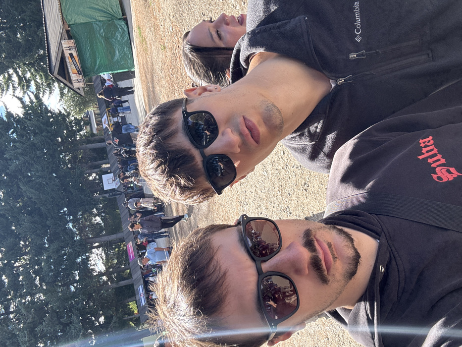 A selfie of three people outside on a sunny day. Two men in sunglasses are at the bottom of the frame, wearing dark-colored jackets. One jacket features "Columbia" on the sleeve, while the other has red lettering. A woman is positioned behind the men. Behind them, there's a crowd of people, trees, a building covered by a green tarp, and a basketball hoop.