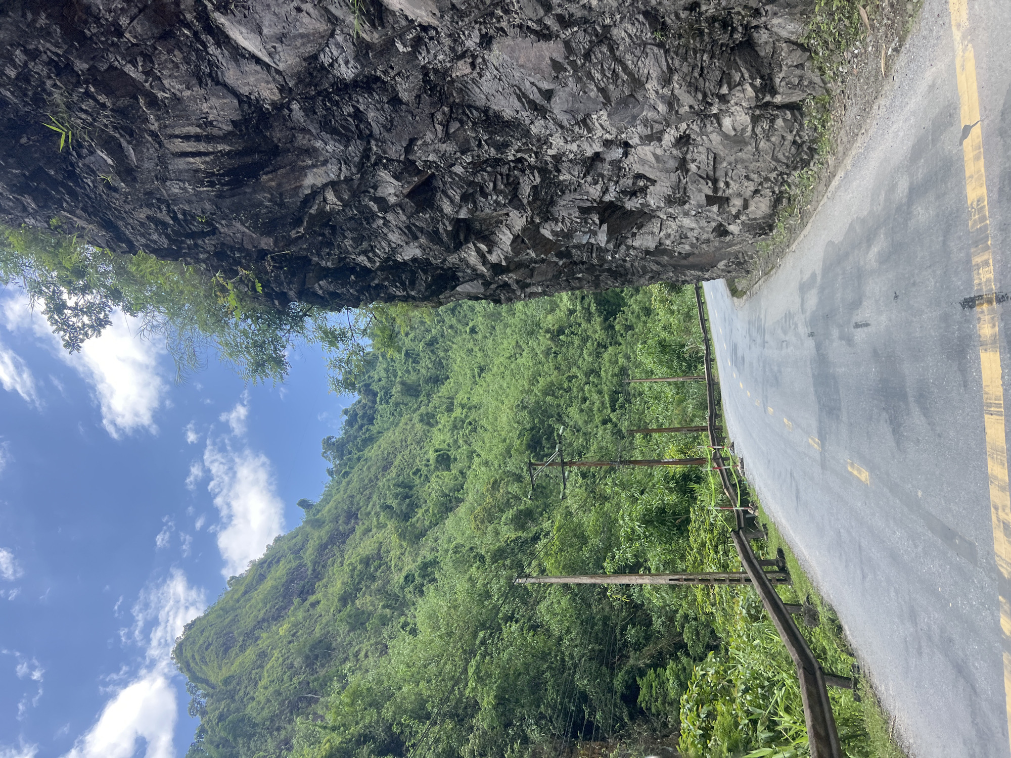 A scenic mountain road curves through a lush, green landscape under a bright blue sky with scattered white clouds. The road surface is asphalt, marked with yellow lines. To the right, a steep, rocky cliff face rises dramatically. On the left, the mountainside is densely covered with vegetation, including bamboo, and dotted with utility poles and power lines. A guardrail runs along the edge of the road on the left. The scene depicts a rural, mountainous environment, likely in Minh Tân Commune, Vietnam, characterized by its natural beauty and infrastructure for transportation.