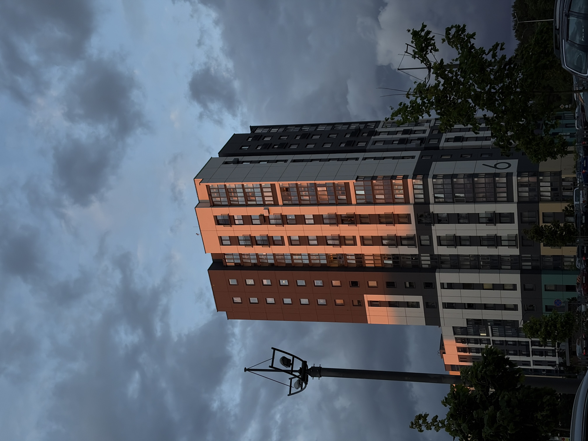The image depicts a tall, modern apartment building in Бараўлянскі сельскі Савет, Belarus, under a cloudy sky. The building has a varied color scheme, with sections in brown, white, gray, and black. The setting sun casts a warm glow on the building's facade. A streetlight is visible in the foreground. Trees and parked cars can be seen along the side of the building. The sky is filled with dark clouds.