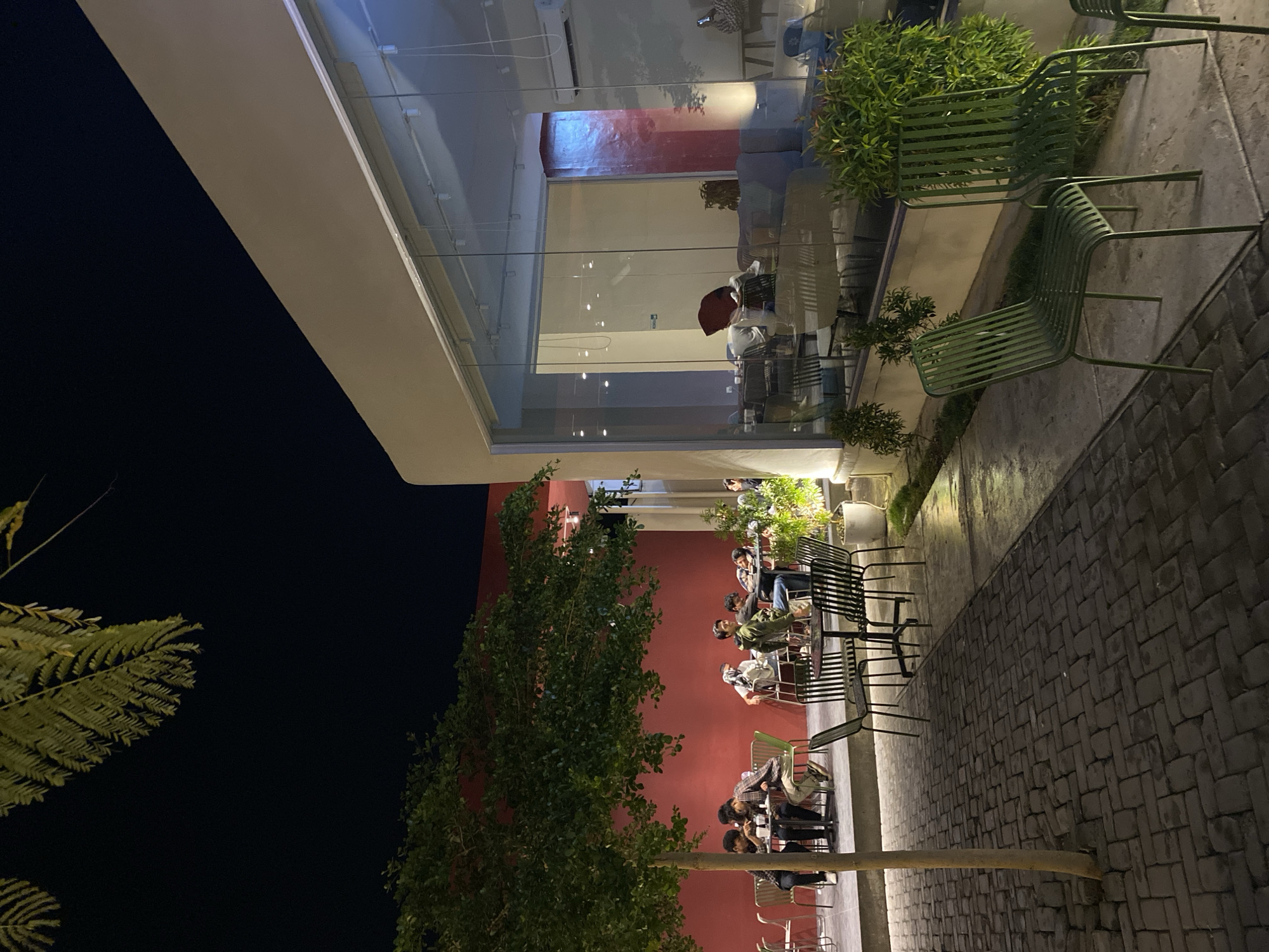 The media depicts a nighttime scene of a modern cafe or restaurant, featuring both indoor and outdoor seating areas. The dark sky indicates it is evening.
Inside the building, visible through large glass walls, people are seated at tables. One individual wearing a red head covering is prominently visible. The interior space appears well-lit with artificial light, revealing an air conditioning unit and some furniture.
Outdoors, several groups of people are gathered at tables, primarily men, engaged in dining or socializing. Some are looking at their phones or conversing. The outdoor area is bordered by a striking red wall and lush green trees and bushes, providing a natural contrast to the built environment. Various metal chairs and tables, including some distinct green and black designs, are arranged on a dark, herringbone-patterned brick pavement. Artificial lighting illuminates the outdoor seating and walking areas, creating bright spots and casting subtle shadows.
The atmosphere appears relaxed and social, typical of an evening gathering spot. The architecture suggests a contemporary design with clean lines and extensive use of glass. No visible text can be discerned in the image. The setting is in Lhokseumawe, Indonesia.