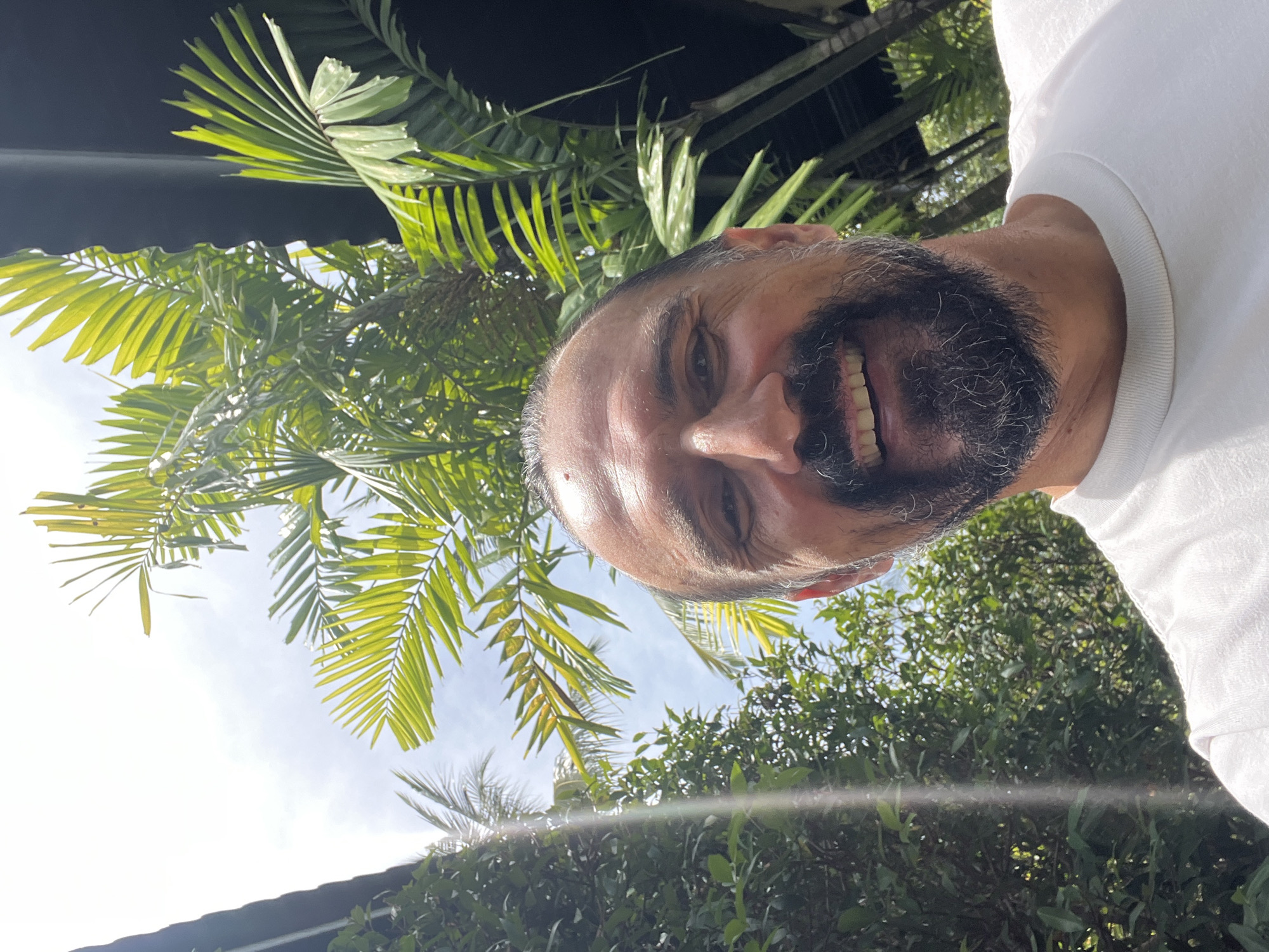 The image is a close-up selfie of a middle-aged man with a salt-and-pepper beard and mustache, wearing a white t-shirt. He is smiling broadly, revealing his teeth. The background consists of lush green foliage, including palm trees, and glimpses of a bright, overcast sky. The presence of palm trees and general vegetation may indicate a tropical outdoor environment. The location, บ้านบางรักษ์, Thailand, is consistent with a tropical climate.