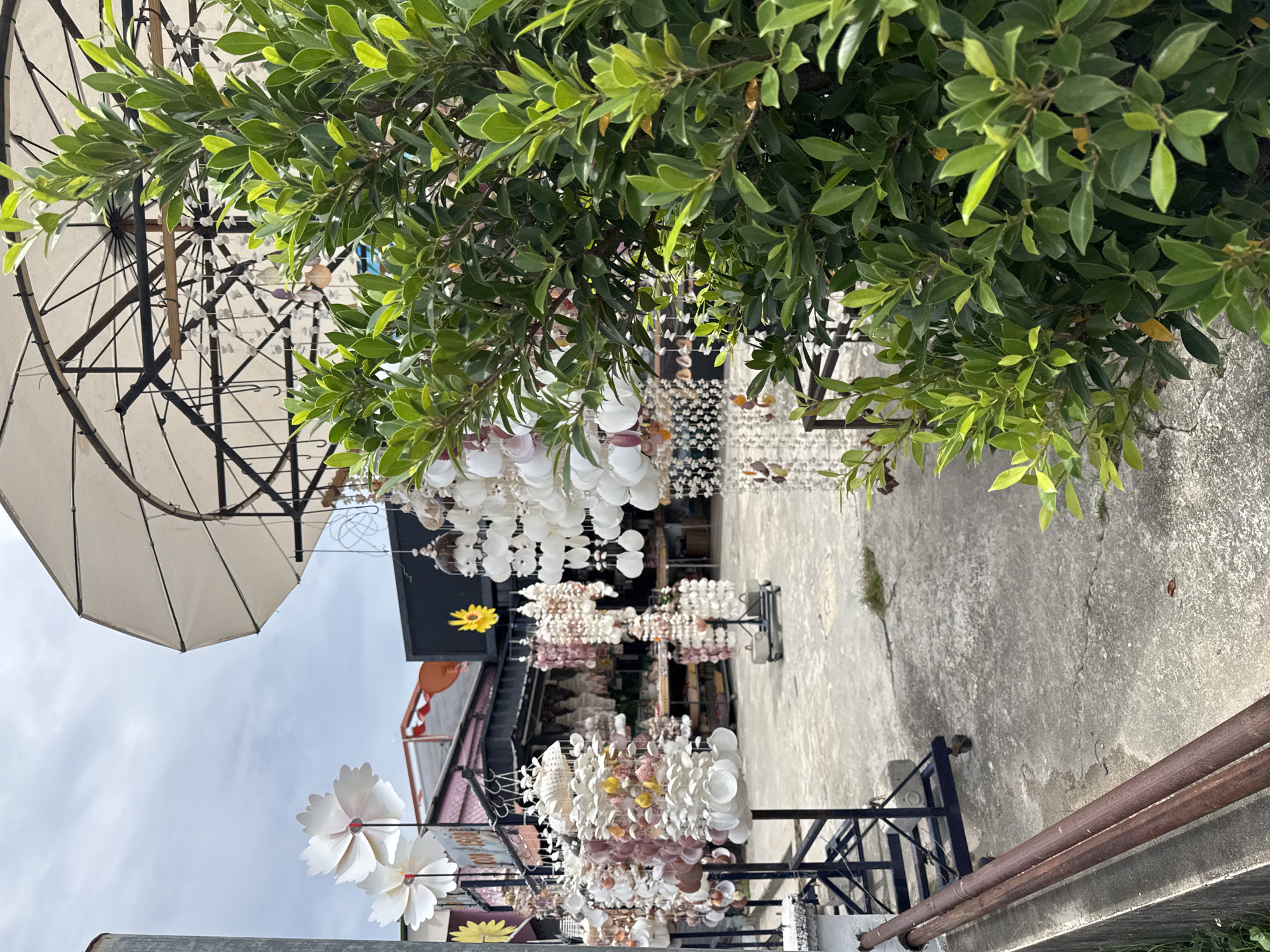A marketplace in Rawai, Thailand, is shown in this medium-angle, outdoor shot. The foreground is dominated by a lush green shrub, obscuring some of the scene. Behind it, a variety of shell ornaments hang from displays, catching the light. Some of these ornaments are delicate and white, while others have hints of pink and yellow. In the background, buildings with sloped roofs are visible, and a large, light-colored umbrella shades a portion of the area. The ground is paved with concrete, showing some cracks and patches of dry grass. A metal pole and some rusty pipes are also present in the foreground, suggesting an industrial or service area adjacent to the market. The sky is overcast. No people are visible in the frame.