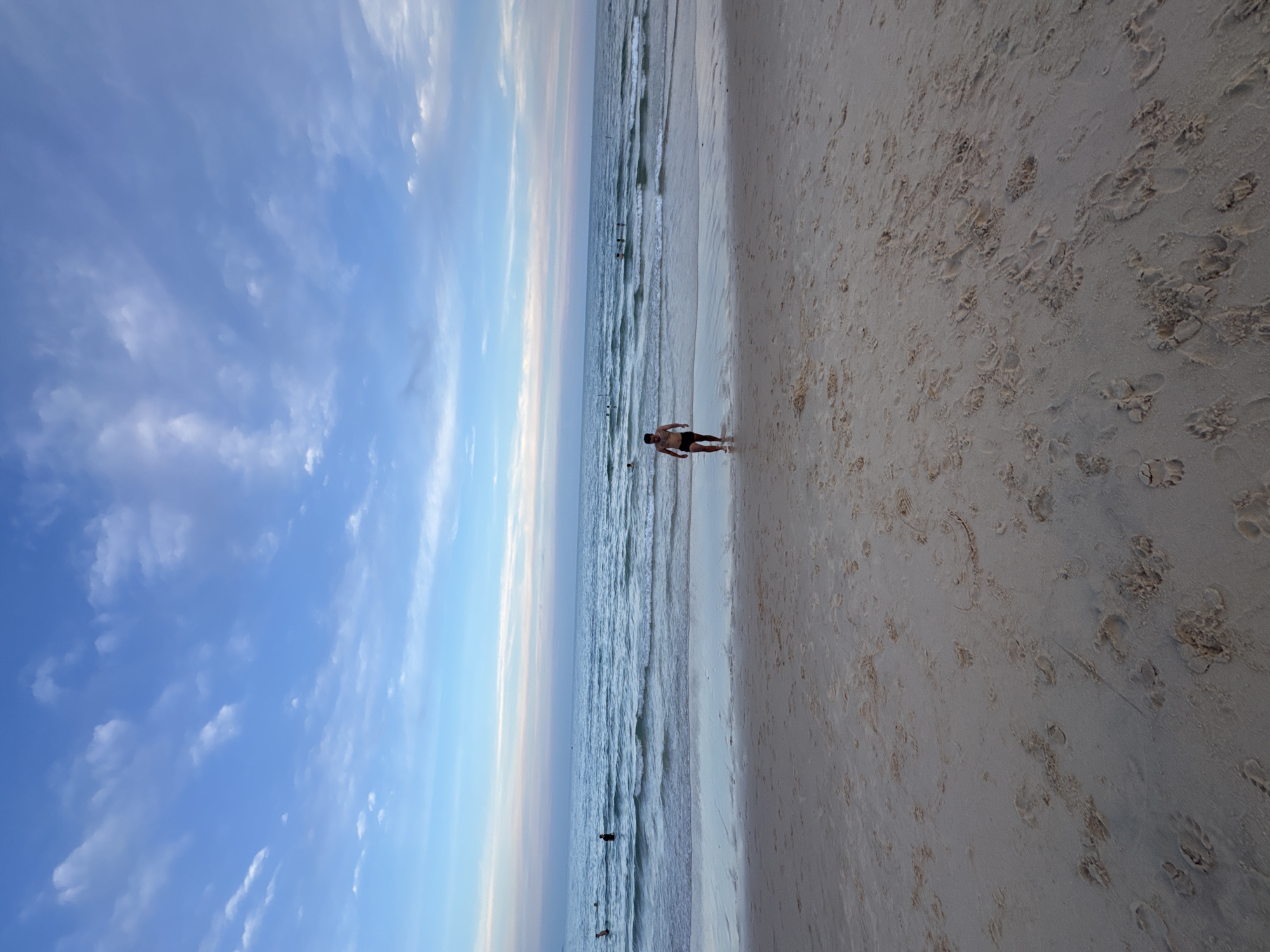 A man in black swim shorts walks towards the ocean from a sandy beach. The man is shirtless and his back is to the camera. The water is calm with small waves, and there are other people in the distance swimming. The sky is blue with scattered white clouds. The beach is wet and sandy with footprints. The scene appears to be at a beach in Phu Thuy Ward, Vietnam.
