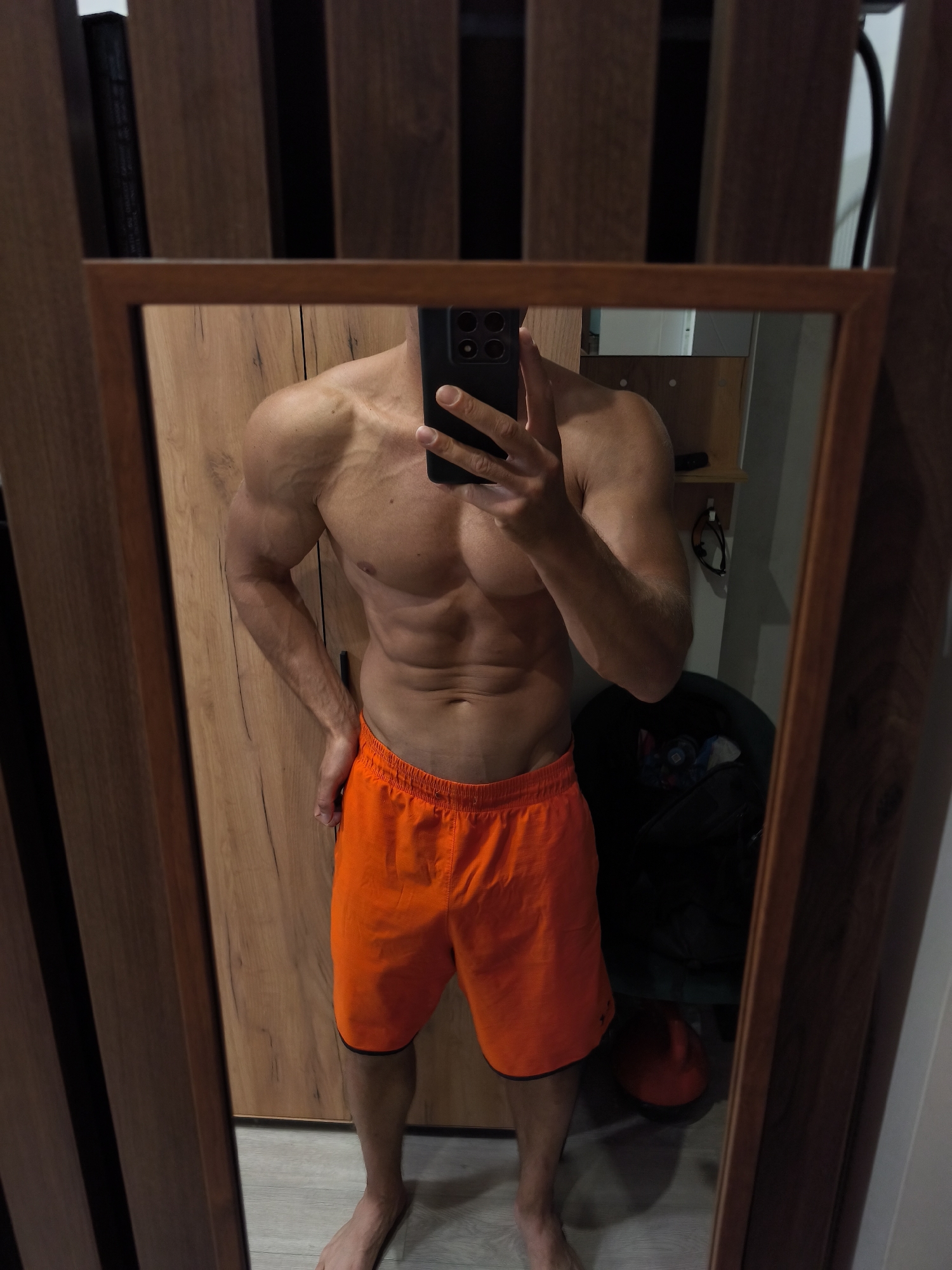 The image shows a person taking a mirror selfie in what appears to be a residential setting. The person is shirtless, wearing orange shorts, and has a muscular physique. They are holding a smartphone in their right hand to capture the reflection. The background includes a wooden wardrobe and a mirror with a wooden frame. There are also some objects visible in the reflection, such as a bag and a red object. The floor is tiled. The scene is indoors, and there is no indication of the specific location within Moscow, Russia.