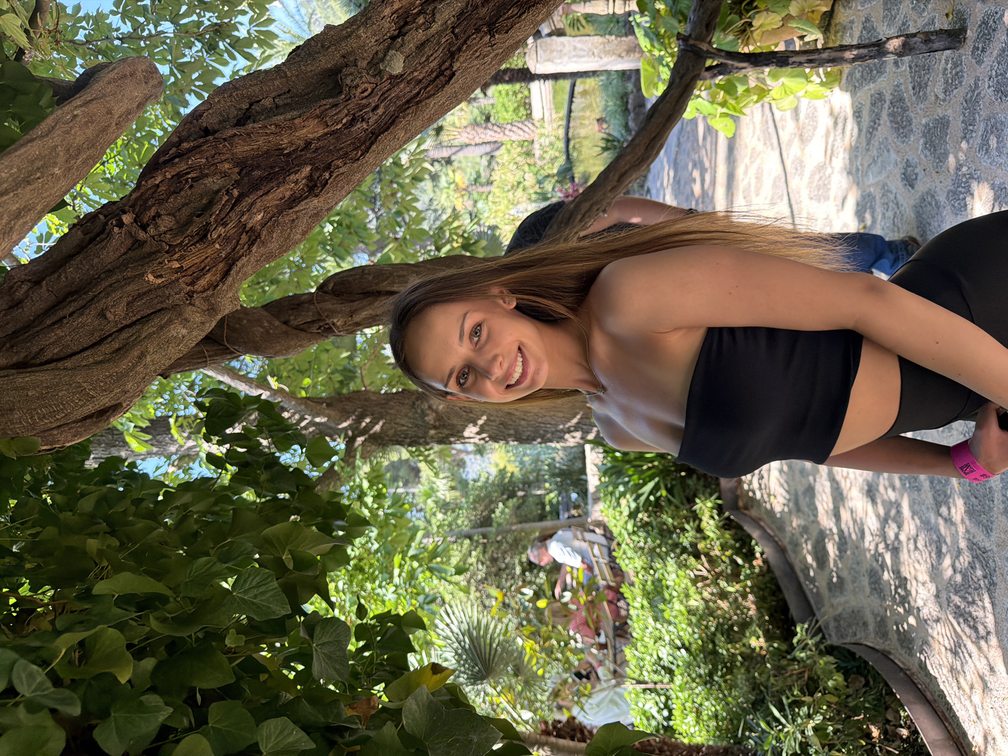 A medium-shot photo shows a woman in a black bandeau top and black leggings smiling and looking towards the camera. She appears to be standing near a tree with thick, gnarled branches, surrounded by lush vegetation. In the background, there is a glimpse of a garden scene with stone pathways and other people. The location may be in Bunyola, Spain, given the presence of stone, a garden, and local vegetation.