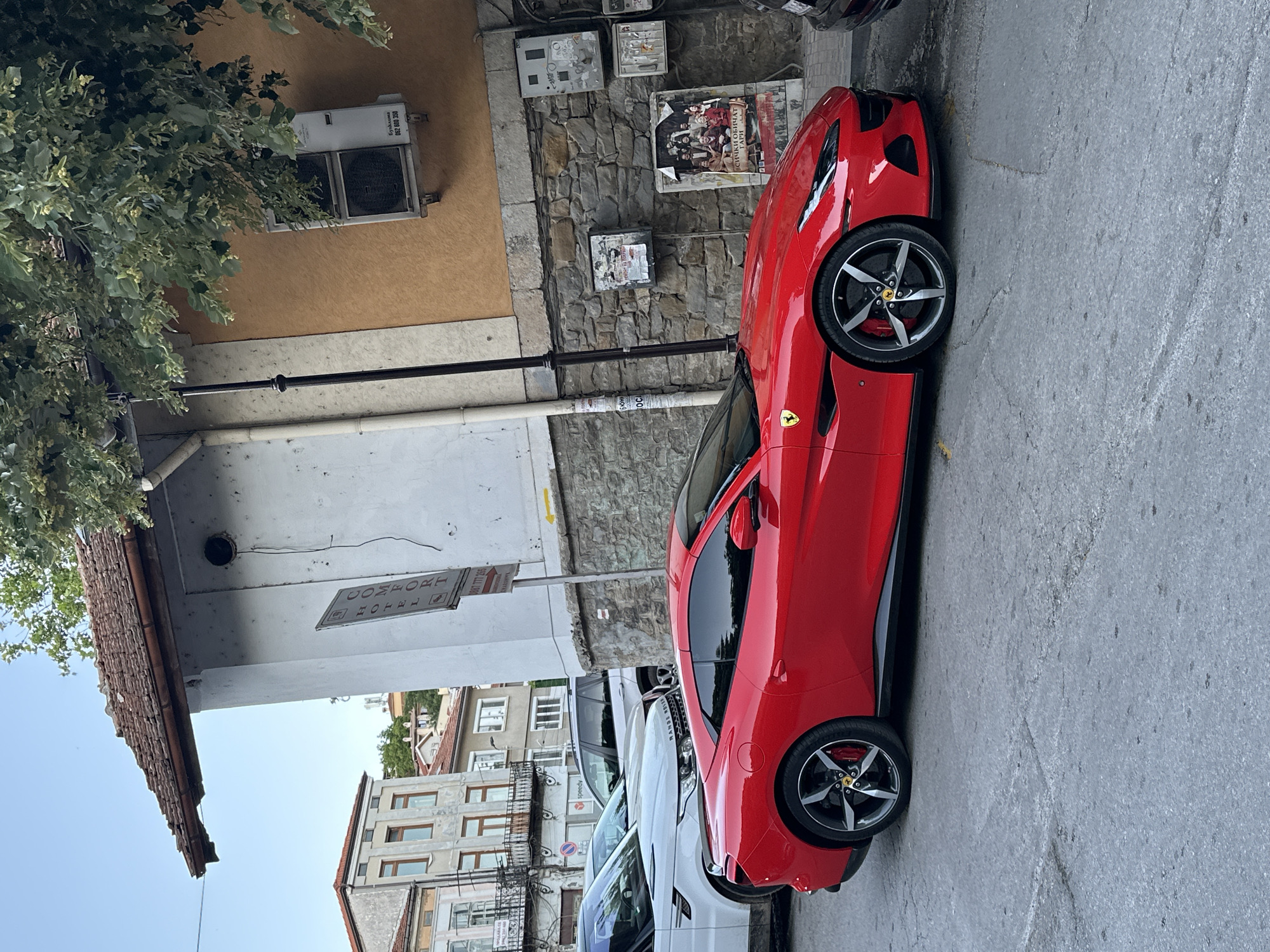 A bright red Ferrari Portofino is parked on a cobblestone street in Veliko Tarnovo, Bulgaria. The car is in the foreground, angled towards the viewer's left. To the left of the Ferrari, a white Range Rover is partially visible. Behind the cars, a stone building with a brown tiled roof is shown. The building has a white facade on the left and an orange facade on the right, with some posters and an air conditioning unit attached to the walls. The sky above is clear blue with some greenery from a tree hanging into the frame. The street appears to be in a historical part of the city.