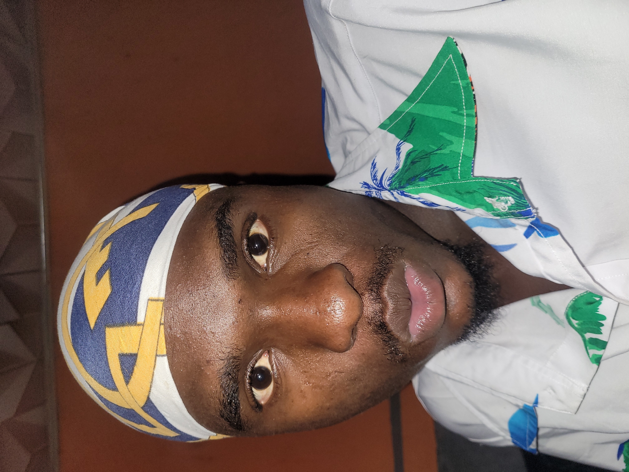 A medium close-up shot shows a young Black man wearing a blue and yellow patterned durag, a white shirt with a green and blue palm tree print, and a goatee. He is looking directly at the camera. The background is a solid reddish-brown wall and a textured ceiling. The image appears to have been taken in Bukuru, Nigeria, based on the provided context.