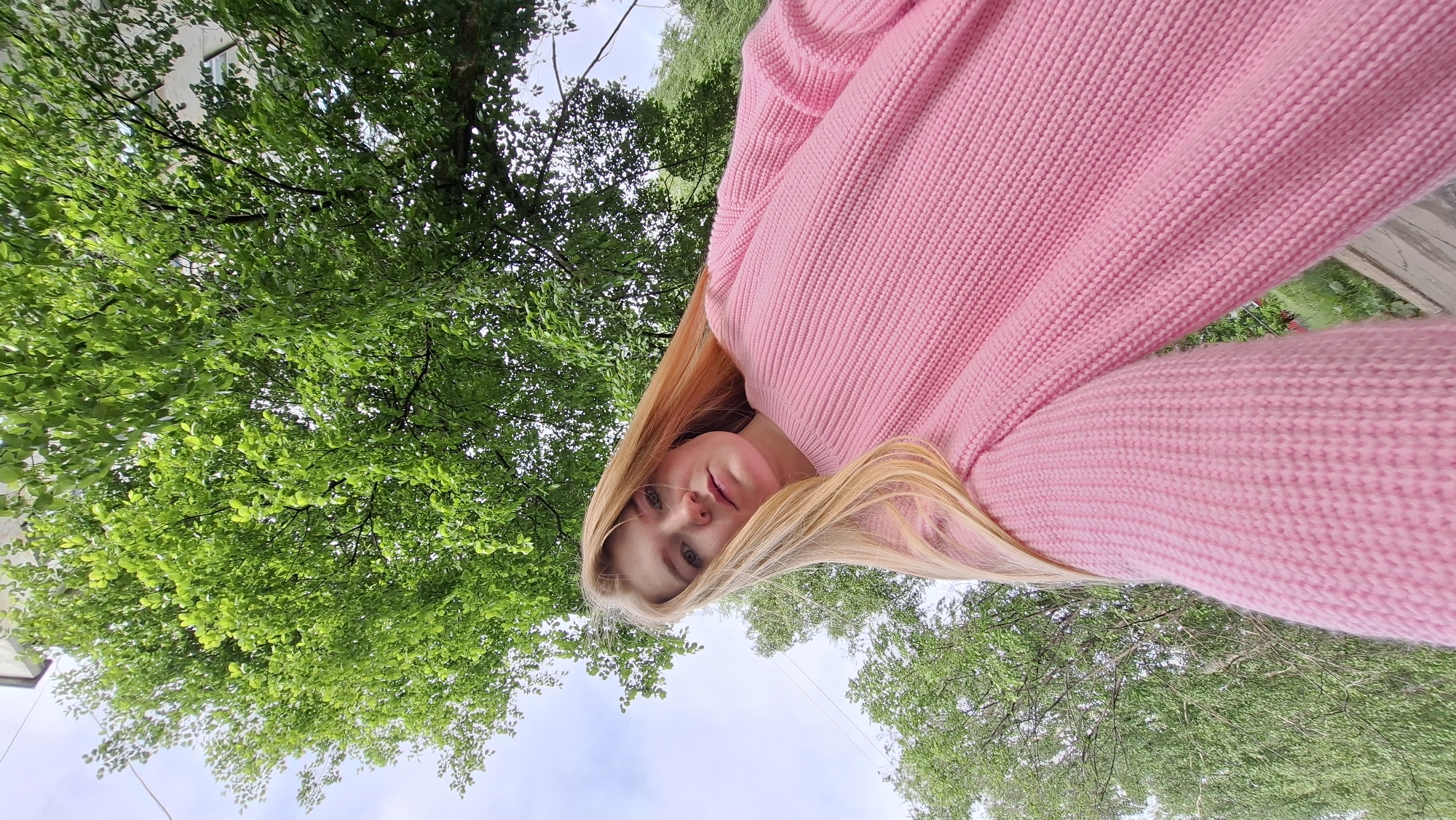 A low-angle, tilted outdoor portrait photograph features a young woman with long, straight blonde hair and a neutral expression, looking directly into the camera. She is wearing a bright pink, ribbed knit sweater. Her face is positioned in the lower-middle section of the frame, and her hair falls around her shoulders.
The background is dominated by the dense, vibrant green foliage of deciduous trees, suggesting a lush outdoor environment, likely during spring or summer. Above and through the tree canopy, a bright, partially overcast sky is visible. A faint, light-colored building or structure can be discerned through the leaves in the upper left portion of the background. The natural lighting indicates it is daytime. No visible text is present in the image. This photograph was taken in Chaikovsky, Russia.