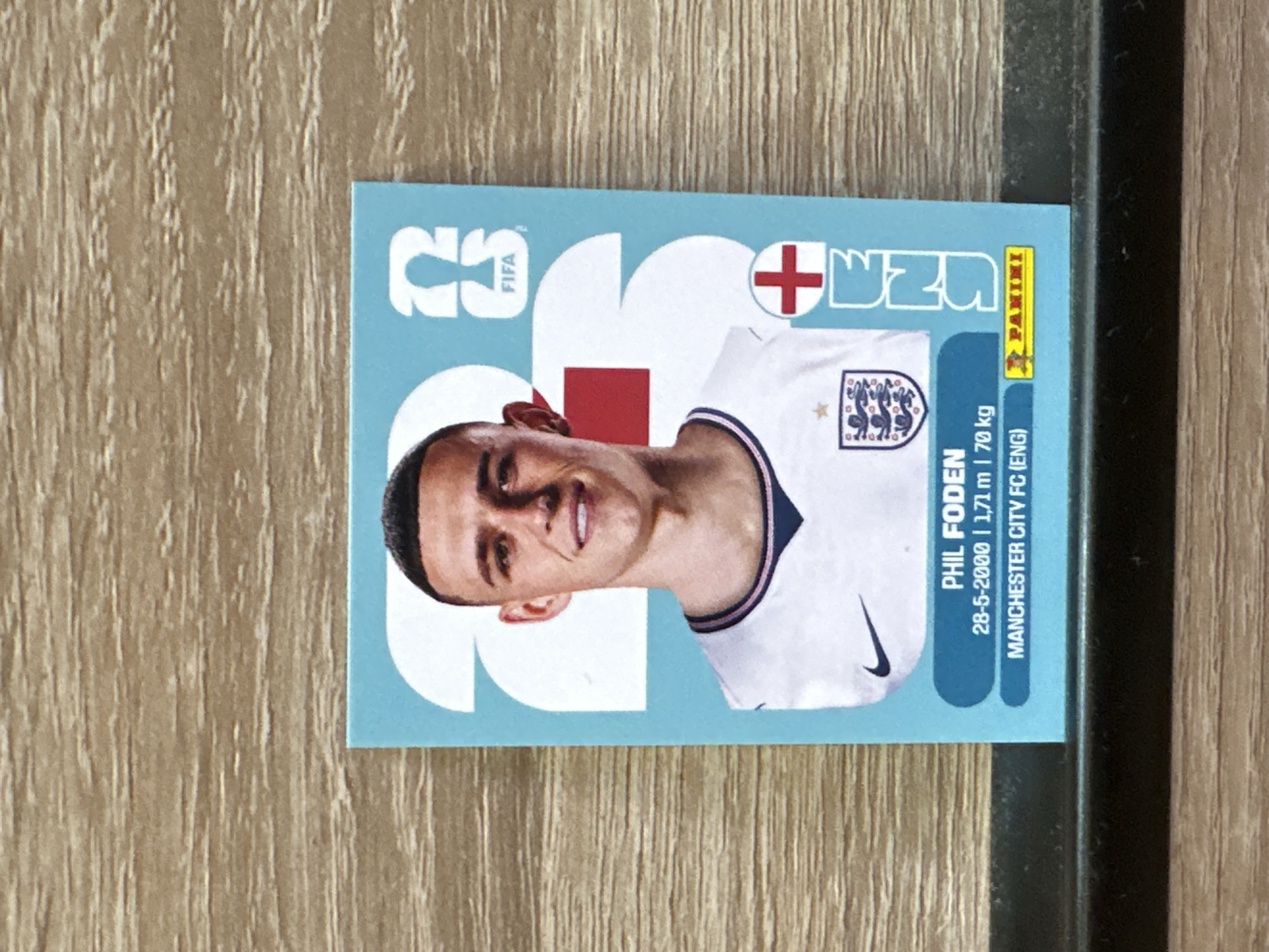 An England football sticker featuring Phil Foden is displayed on a wooden surface. The sticker has a light blue background with a large white graphic of the number '2' and part of a '3'. Phil Foden is depicted in the center wearing an England jersey. Below his image, text reads "PHIL FODEN", "28-5-2000 | 1,71 m | 70 kg", and "MANCHESTER CITY FC (ENG)". The Panini logo is visible in the bottom right corner. The scene is static with no actions or interactions occurring. The environment is a close-up view of the sticker on a textured wooden surface. No elements in the image suggest the city of Prisovo, Bulgaria.