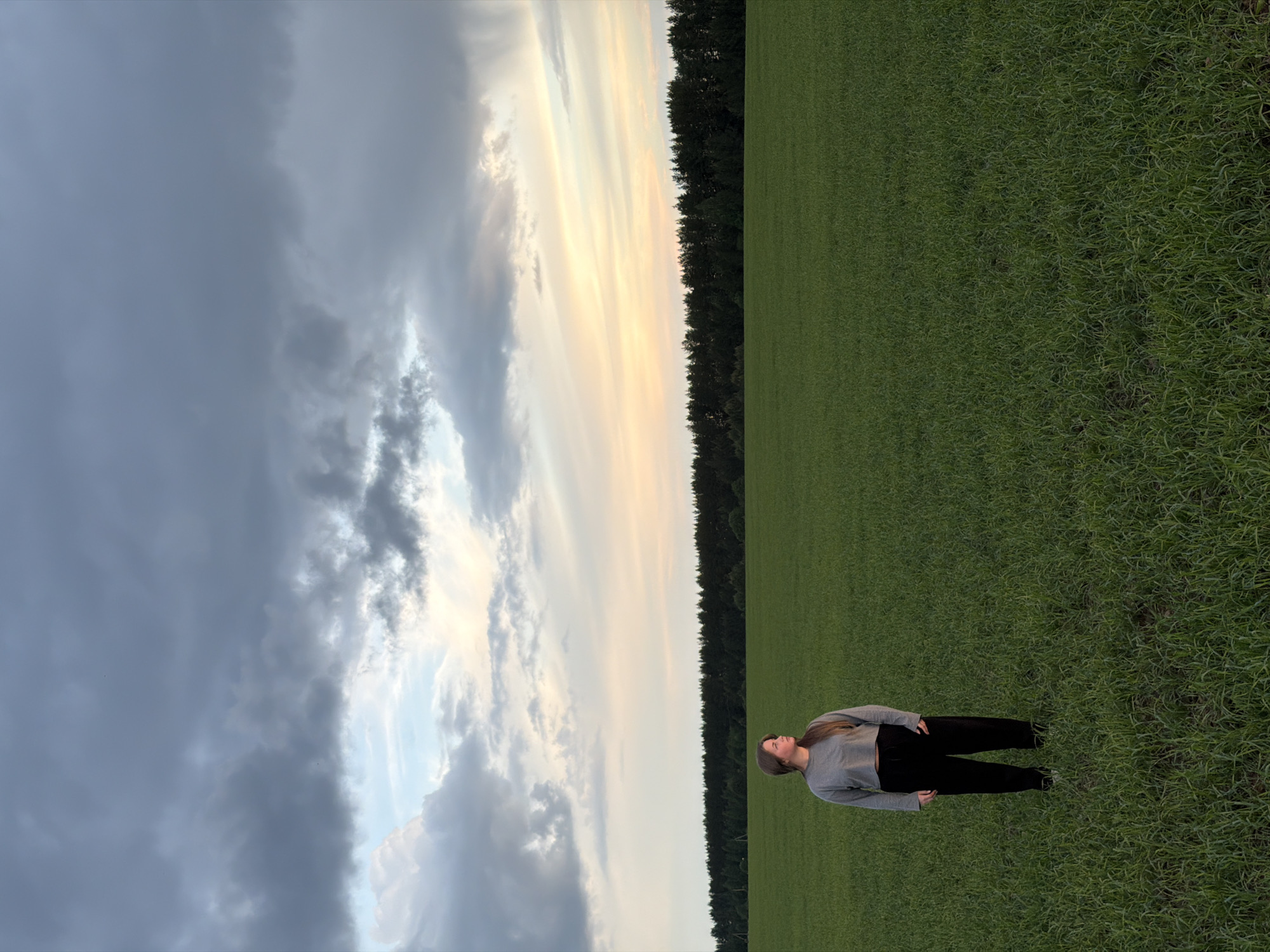 A woman lies on a grassy field on the outskirts of Фоки, Russia. The sky above is partially obscured by dark, dramatic clouds, while the setting sun casts an ethereal glow on the horizon. A line of trees separates the field from the sky, and the woman lies perpendicular to this line, looking up at the sky.