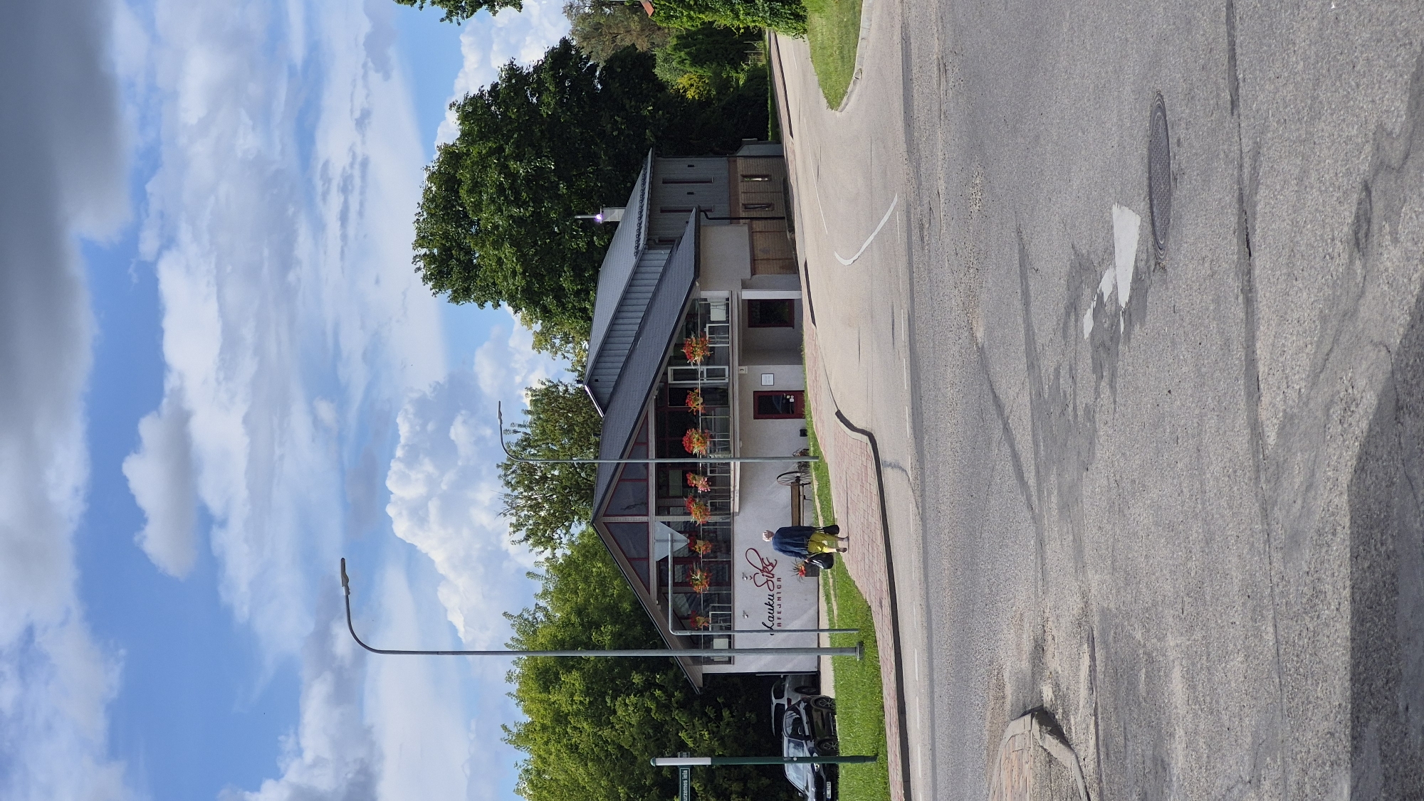 The image shows a modern building with a prominent sign that reads "Lauku Sēta". The building has large windows and a balcony with flower planters. A person wearing yellow pants and a blue jacket is walking away from the camera, carrying two bags. The building appears to be a restaurant or cafe. It is situated on a street corner with a paved road and a sidewalk. The sky is partly cloudy, and there are trees in the background. The street sign on the left side of the image reads "Lielā iela". Lielā iela is the main street in Jēkabpils, Latvia.