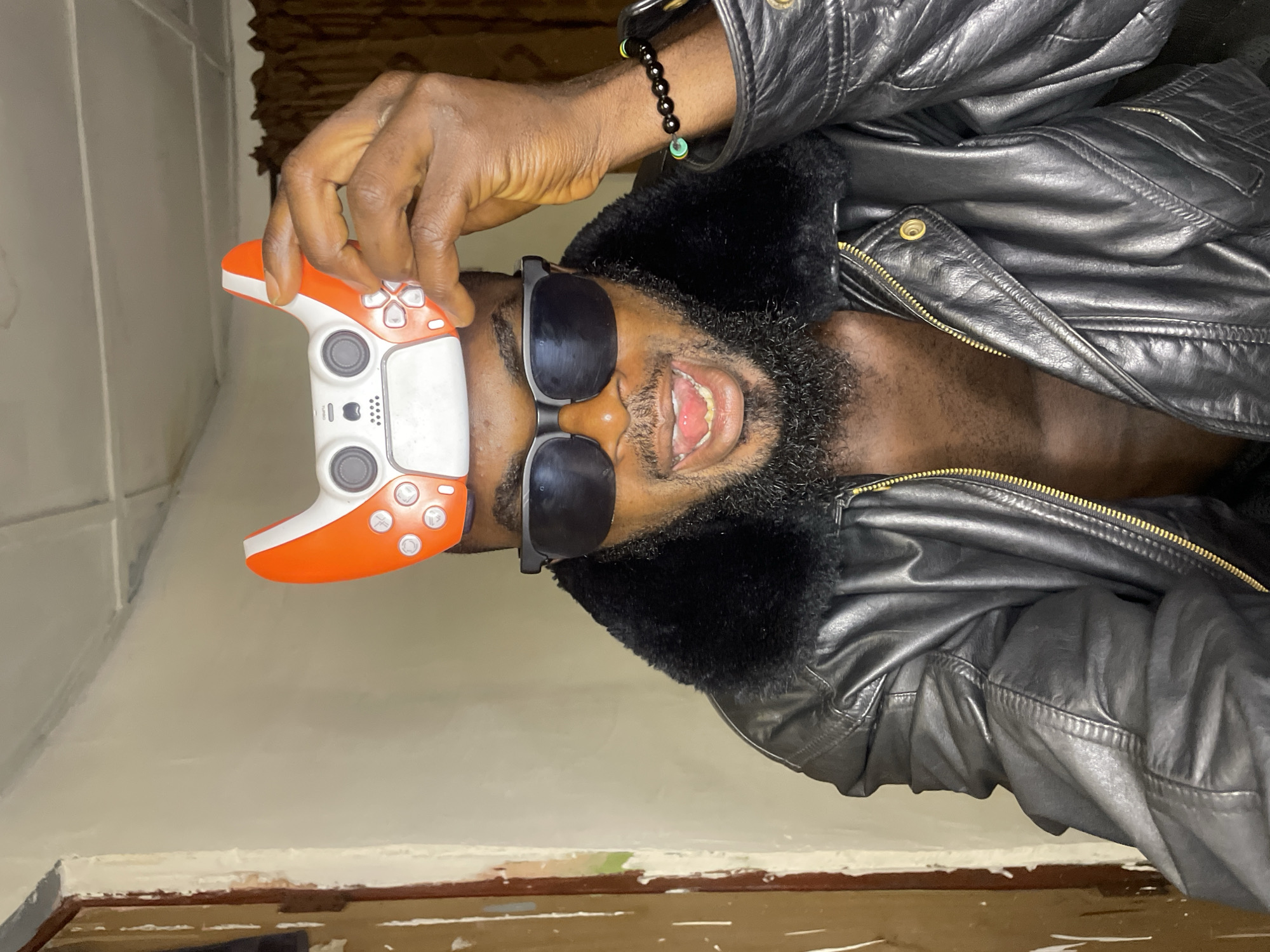 A Black man is facing upwards, wearing black sunglasses and a black leather jacket with a dark fur collar. He holds a white and orange video game controller above his head with his right hand. He has a dark beard and is smiling. A black beaded bracelet is visible on his right wrist. The background is a pale yellow wall with a dark brown curtain visible in the top right corner. There is no indication of the location being in Jos, Nigeria.