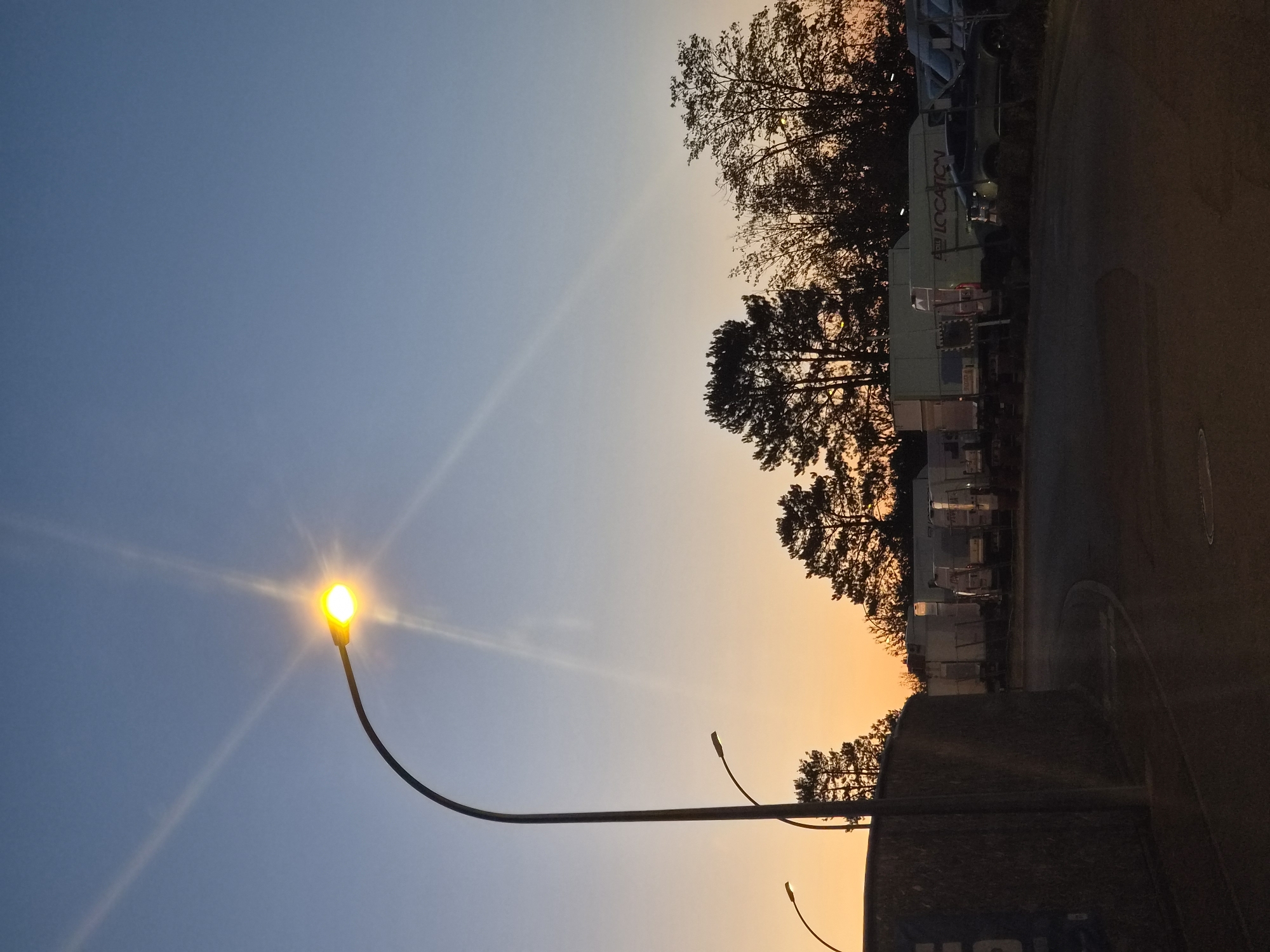The image captures a street scene at dusk in Leudelange, Luxembourg. A tall lamppost with its light illuminated dominates the left side of the frame, casting a bright glow and creating light streaks against the gradient sky, which transitions from a deep blue at the top to a warm orange near the horizon. In the mid-ground, several parked vehicles and trailers are visible, some with their lights on. Silhouetted trees line the horizon behind the vehicles. A dark-colored car is parked in front of a white trailer with "LOCATION" written on its side. The foreground shows a dark asphalt road curving slightly. No people are visible in the scene.