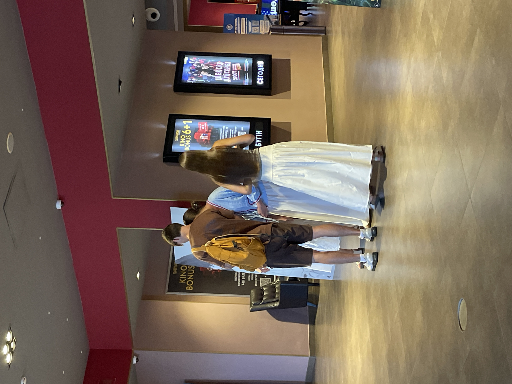 This image shows three individuals standing in what appears to be the interior of a cinema lobby. The lighting is dim, and the walls are a muted orange and pink.
In the foreground, a young man wearing a brown t-shirt and shorts, with a yellow backpack slung over his shoulder, stands with his back to the camera. To his right, a woman in a light blue, traditional-looking dress with white embroidery stands with her side to the camera. Further to the right, another woman with long dark hair, wearing a gray tank top and a long white skirt, stands with her back to the camera.
Behind the people, there are several illuminated movie posters displayed on the wall. One poster on the left advertises "KINO BONUS 6+1". Another poster to its right displays a movie title in Cyrillic script that translates to "SEX AND THE CITY". The floor is tiled with large, dark gray tiles. In the mid-ground, a black leather armchair is visible.
The presence of movie posters, typical lobby furniture, and the overall aesthetic suggest a cinema environment. The Cyrillic script on the posters strongly indicates a location within a Russian-speaking region. Considering the prompt specifies Almaty, Kazakhstan, this scene is consistent with a cinema lobby in Almaty. There is no visible indication of the exterior or any specific landmarks that would definitively place it in Almaty.