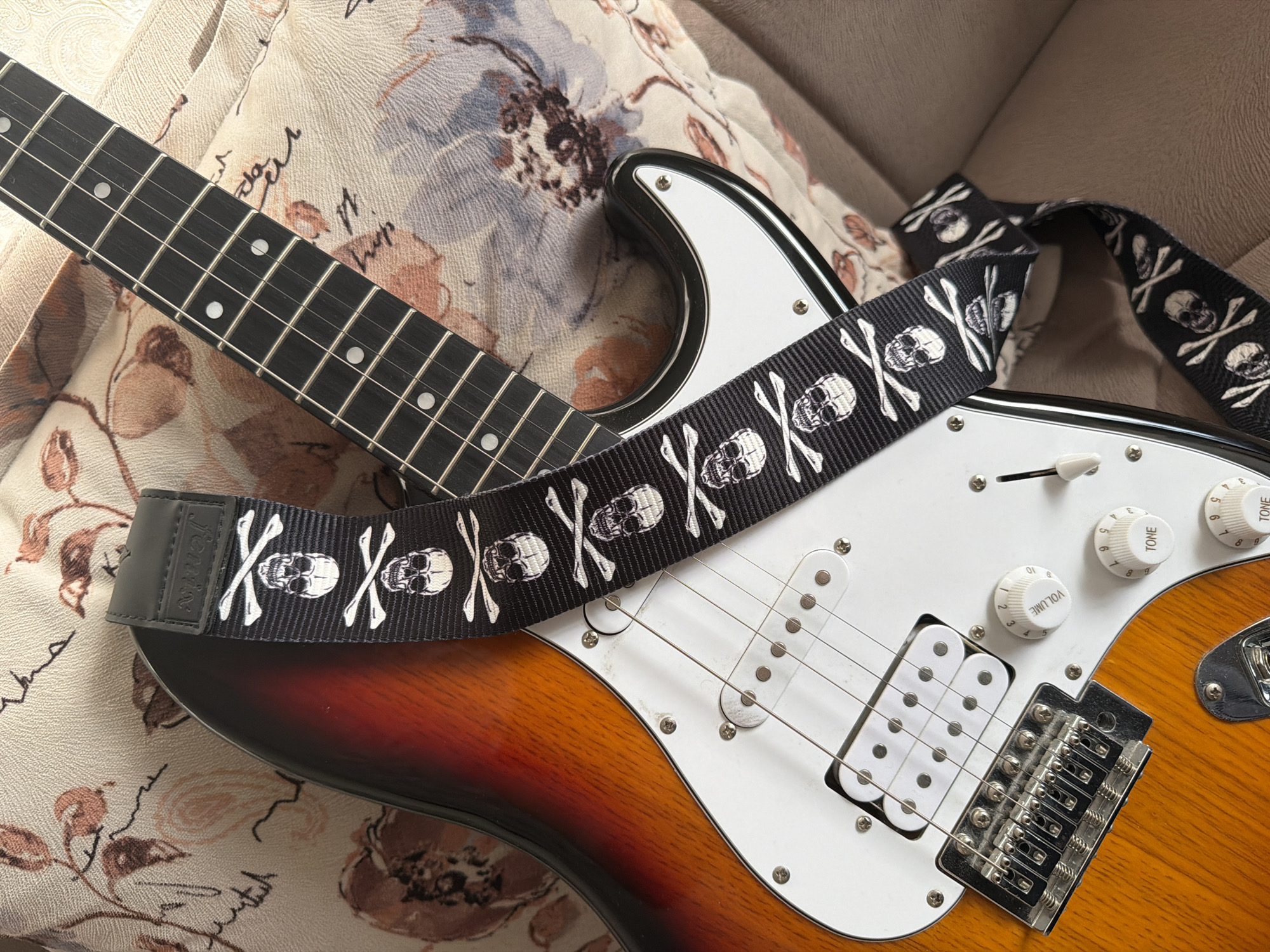 An electric guitar with a skull and crossbones pattern strap rests on a textured fabric surface that appears to be a decorative cushion. The guitar has a sunburst finish on its body, transitioning from orange to red. Visible components include the fretboard with white fret markers, pickups, tuning pegs, volume and tone knobs, a bridge with six strings, and a guitar strap. The strap features a repeating pattern of white skulls and crossbones on a black background. The background fabric has a floral pattern in muted tones of brown, pink, and blue, with some subtle script-like markings. The lighting suggests an indoor setting, possibly a room. There are no people present in the image, and no specific location in Baku City, Azerbaijan can be identified.