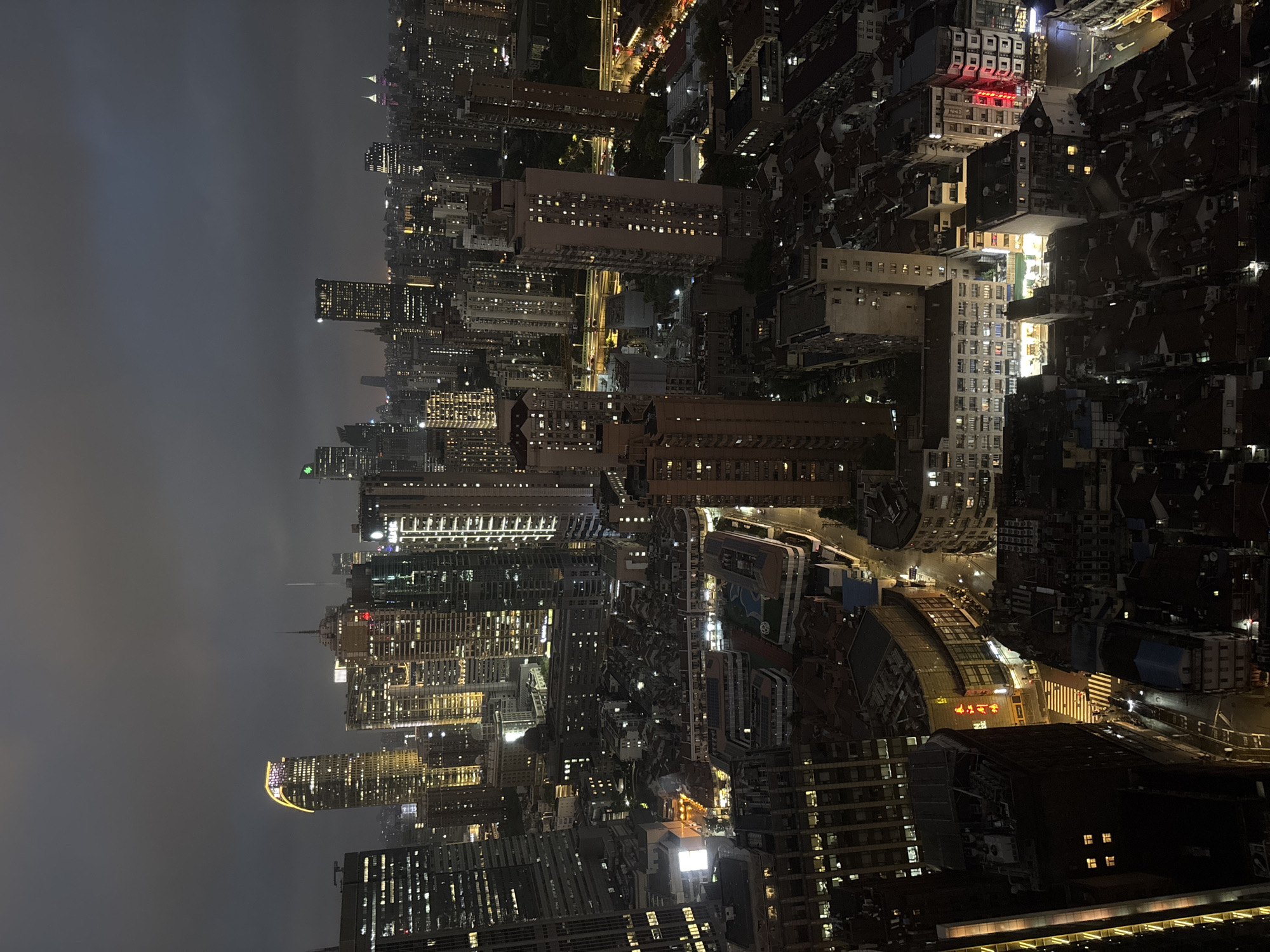 This is an aerial nighttime photograph of the Xintiandi area of Shanghai, China. The image captures a dense urban landscape characterized by a mix of modern skyscrapers and older, lower-rise buildings. The sky is overcast and dark, with only a hint of light breaking through.
Dominating the skyline are several prominent skyscrapers, many of which are illuminated with a warm, golden light, highlighting their architectural features. One building, in the upper left quadrant, has a distinct curved top illuminated with a bright golden band. Other skyscrapers display grids of lit windows.
In the mid-ground and foreground, the architecture shifts to reveal a more traditional urban fabric. This area features numerous smaller buildings with red-tiled roofs, densely packed together. Some of these buildings have illuminated windows, and streetlights cast pools of yellow light on narrow streets. A prominent crosswalk is visible, with a few figures crossing it. There are also indications of commercial activity, with brightly lit signs, one of which appears to be in Chinese characters.
The overall impression is one of a vibrant, bustling city at night, where the old and new coexist. The lighting, the architecture, and the density of the buildings all contribute to a sense of urban dynamism. The image appears to have been taken from a high vantage point, possibly a tall building in the vicinity.